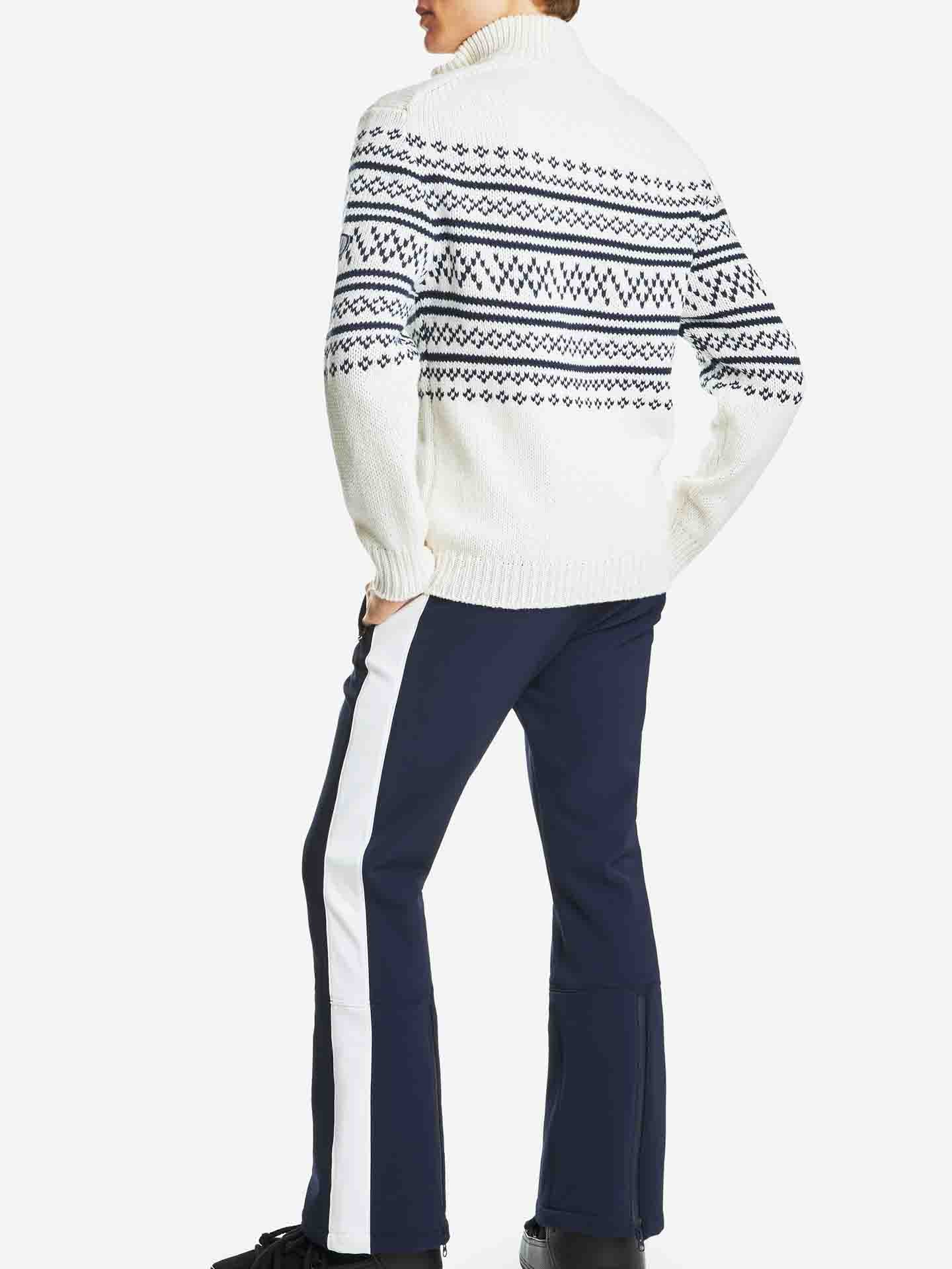 A person is standing with their back to the camera, wearing the We Norwegians Setesdal Zip Up Sweater Men Off White, which features a blue and black Nordic pattern. They pair it with blue pants that have white side stripes and wear black shoes, completing a perfect après ski look. Their left hand is casually in their pocket.