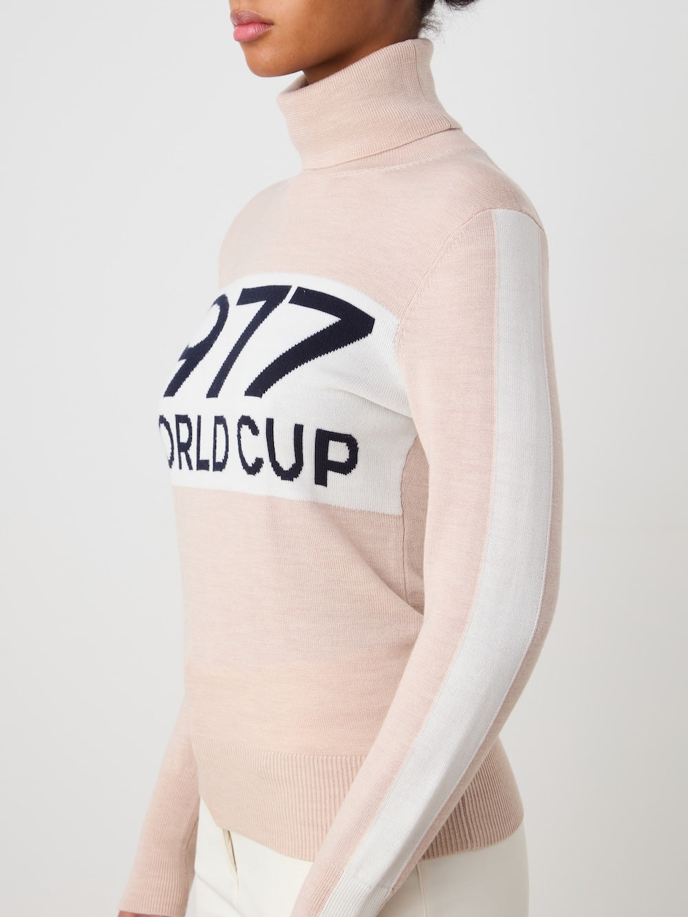 A woman models the We Norwegians World Cup Sweater women - Pre Spring 26, featuring beige knit, white and black "1977 WORLD CUP" chest text, and a bold white sleeve stripe for a modern après-ski vibe against a simple backdrop.