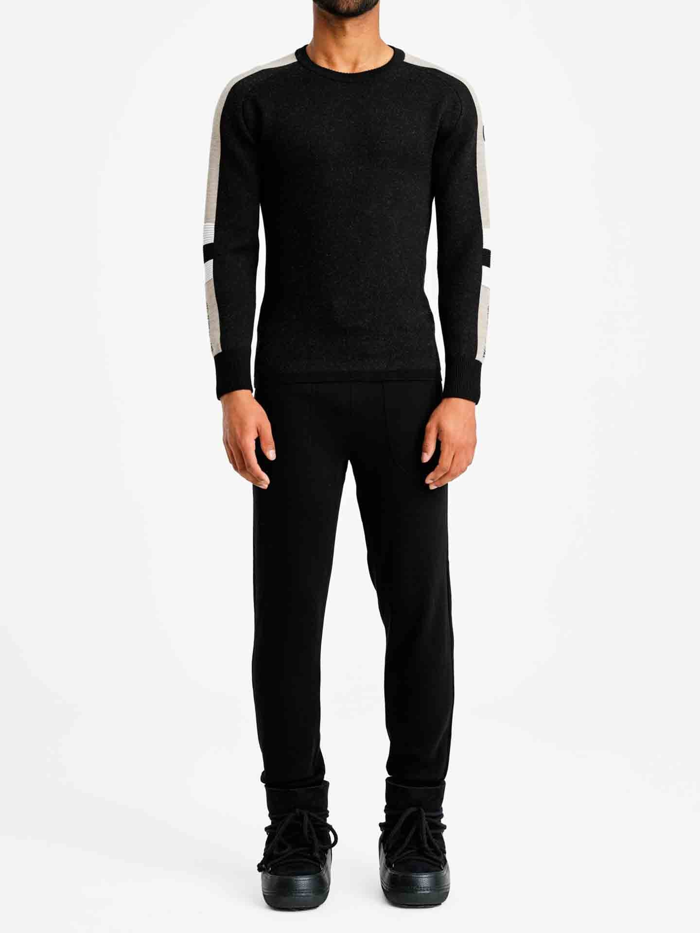 A person stands against a plain white background, wearing the Hafjell Crewneck Men Black from We Norwegians. The sweater features beige and black stripes on the sleeves, paired with black pants and black shoes. The person's arms rest relaxed by their sides, presenting a fitted outfit that appears both casual and stylish.