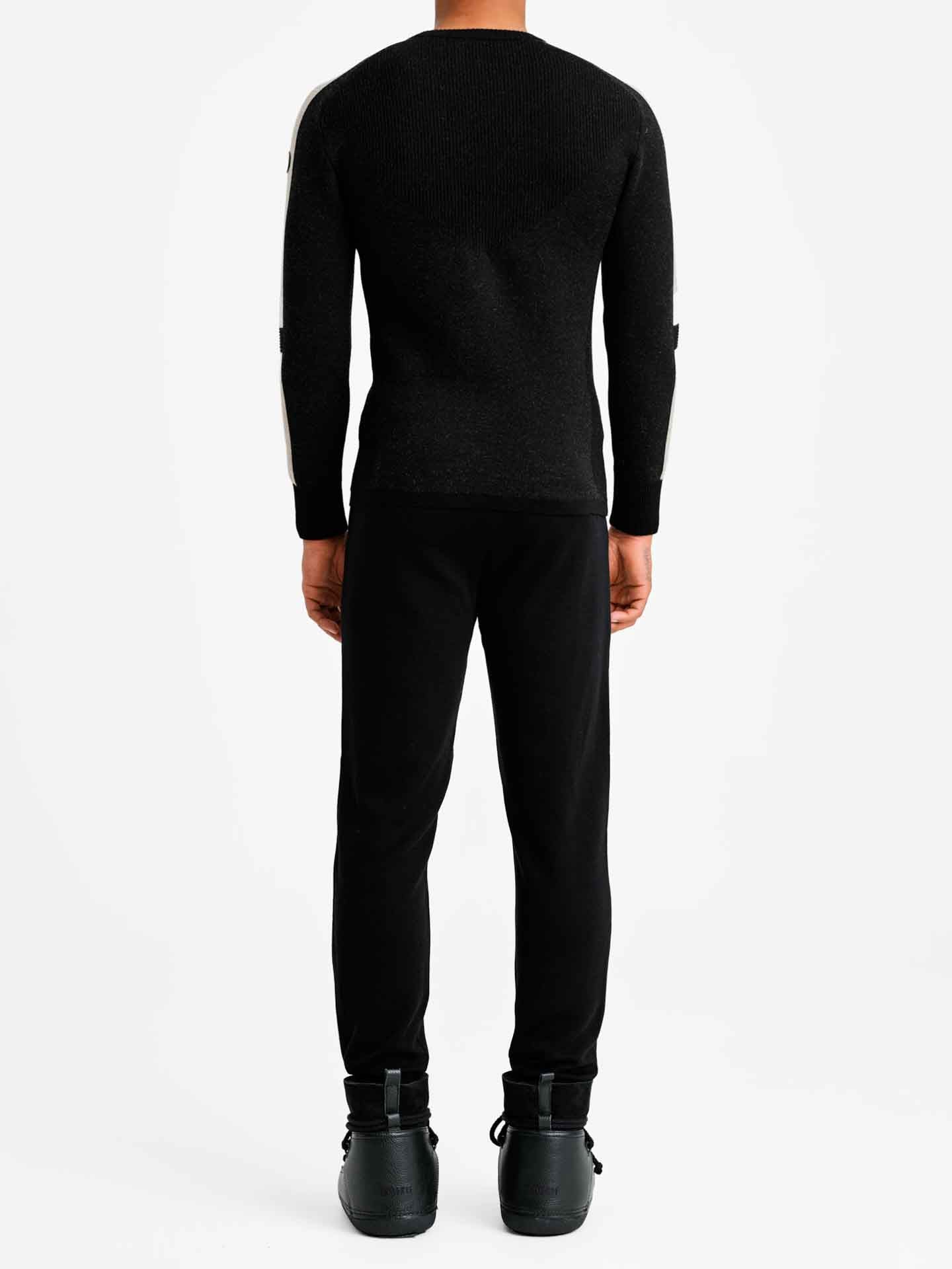 A person stands facing away from the camera, wearing a fitted Hafjell Crewneck Men Black sweater by We Norwegians, along with black pants and black shoes. The background is plain white, emphasizing the silhouette of the individual.