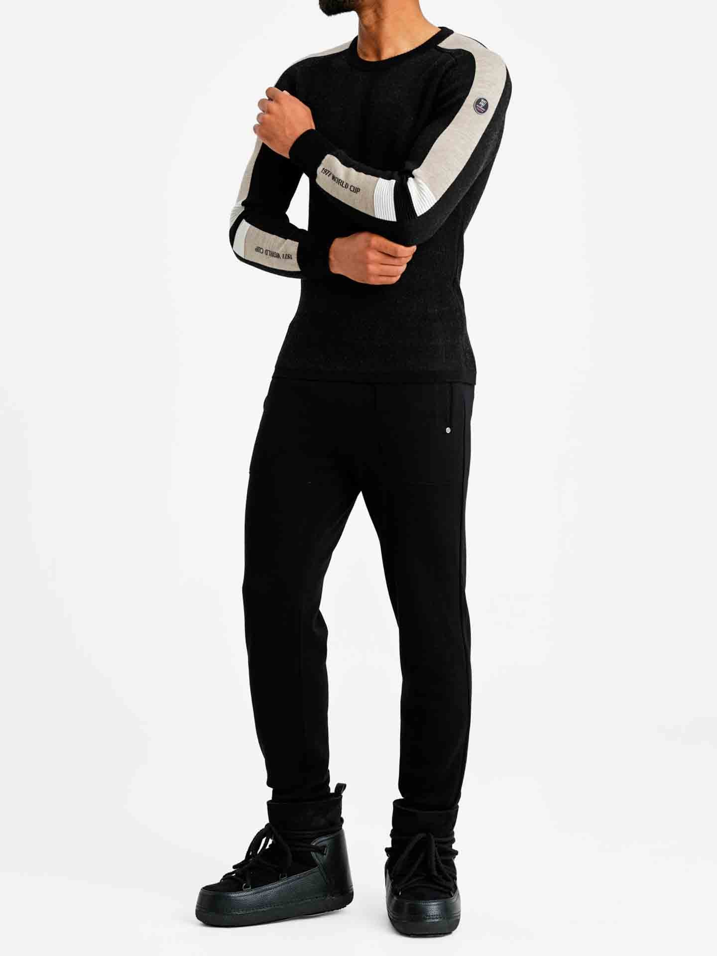 A person standing against a white background is wearing the "Hafjell Crewneck Men Black" by We Norwegians, a black long-sleeve shirt with white stripes on the upper sleeves, reminiscent of classic ski sweaters. Paired with black pants and black high-top sneakers, the individual's face is not visible in the image.