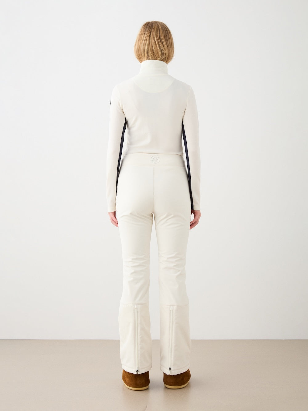 A person with blonde hair wears the We Norwegians Voss Chevron ZipUp Women—an off-white piqué knit turtleneck top and fitted pants with black side panels—paired with brown shoes, standing against a plain white wall in a minimalistic setting.
