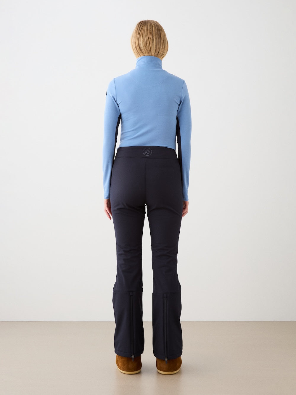 A person with short blonde hair, dressed in the We Norwegians Voss Chevron ZipUp Women, black fitted pants, and brown boots, stands facing away from the camera against a plain background.