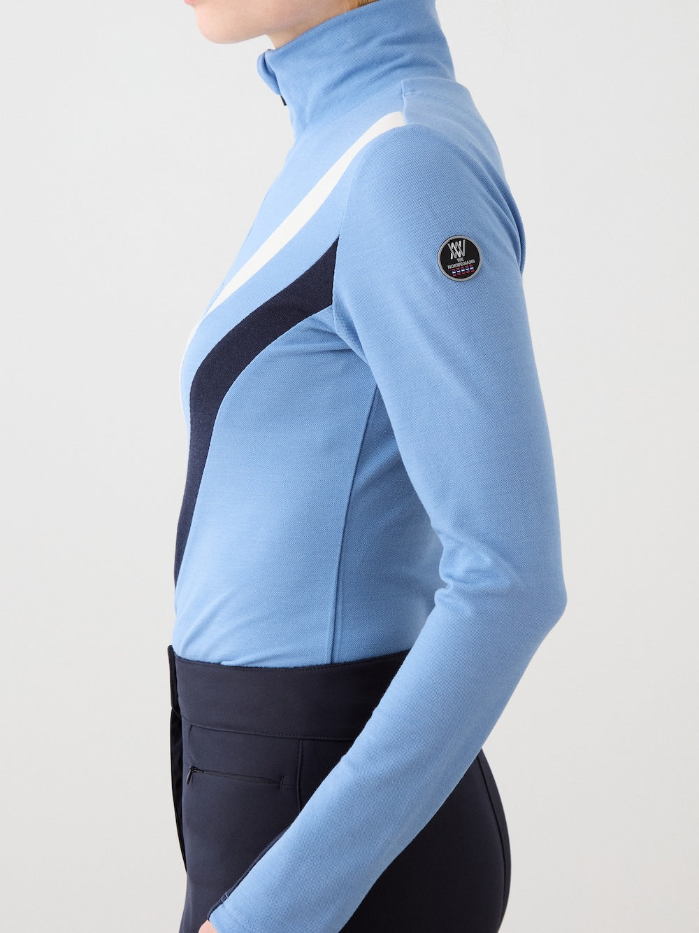 A woman is shown from the side wearing the We Norwegians Voss Chevron ZipUp, a light blue piqué knit base layer with dark blue and white diagonal stripes, high collar, round logo patch on the sleeve, and paired with dark pants.