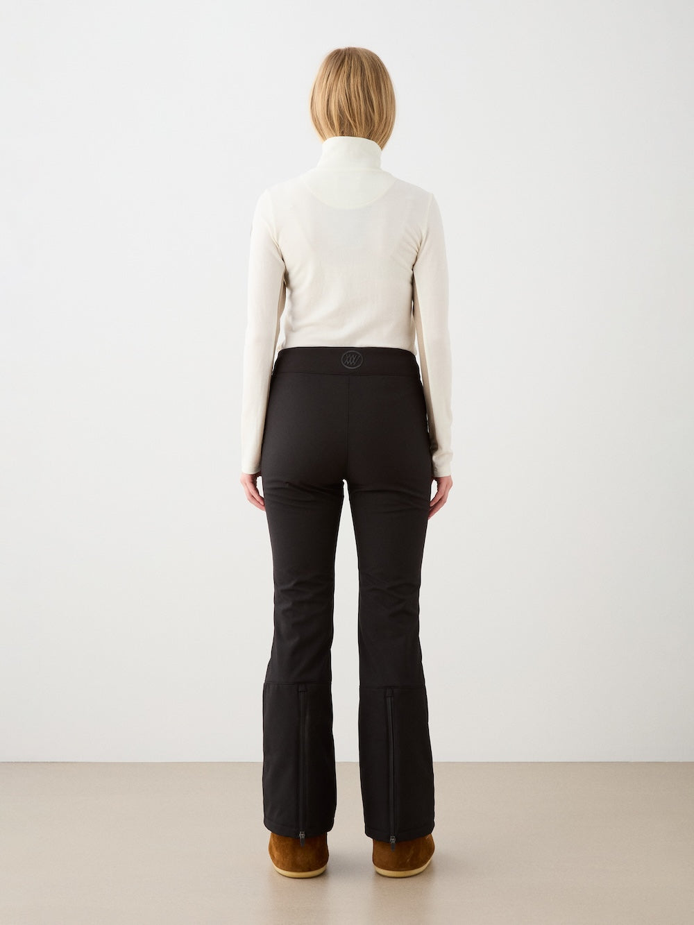 A person with blonde hair wears the We Norwegians Voss Chevron ZipUp, paired with black fitted pants and brown boots, standing with their back to a plain white wall and beige floor.