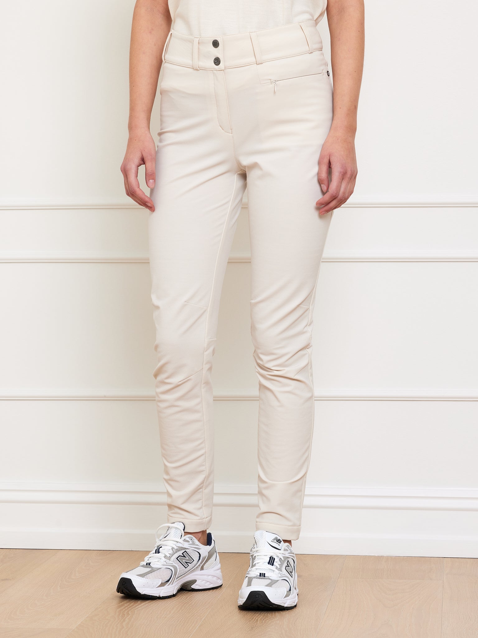 A person wearing We Norwegians' Lista Pant Women Cream stands in front of a cream wall with decorative trim on a light wooden floor. These beige, high-waisted pants feature Swiss Schoeller fabric, double buttons, a front pocket, and 4-way stretch for maximum comfort paired with white sneakers.