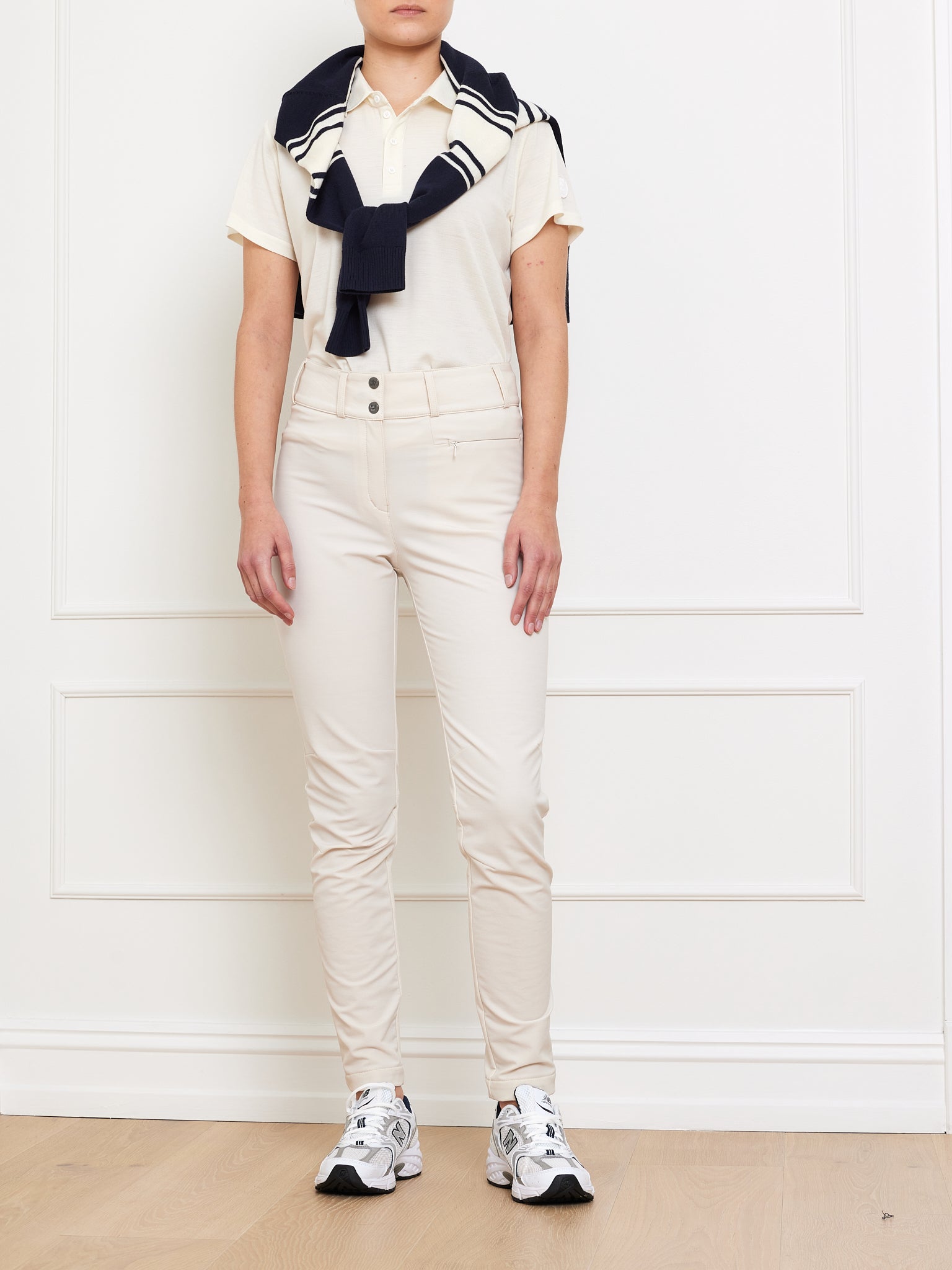 A person stands against a white wall, wearing a light beige polo shirt, We Norwegians Lista Pant in cream made from Swiss Schoeller fabric, and white sneakers. A navy blue sweater is draped over their shoulders, tied at the front. The setting appears minimalist and clean.
