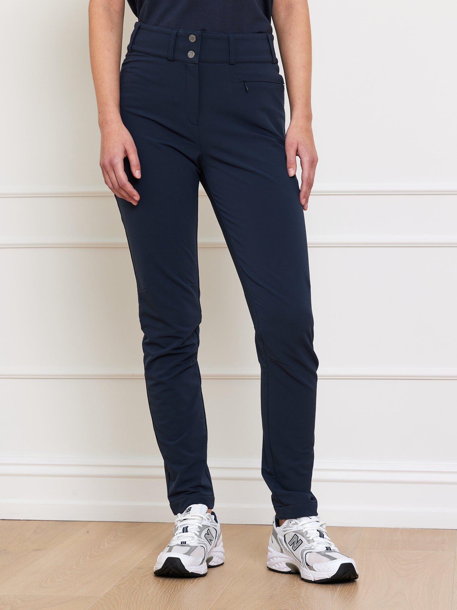 A person in navy Lista Pant Women by We Norwegians, made from Schoeller fabric with high-waist buttons, and white sneakers stands against a white wall with molding. Light wooden flooring completes the scene.