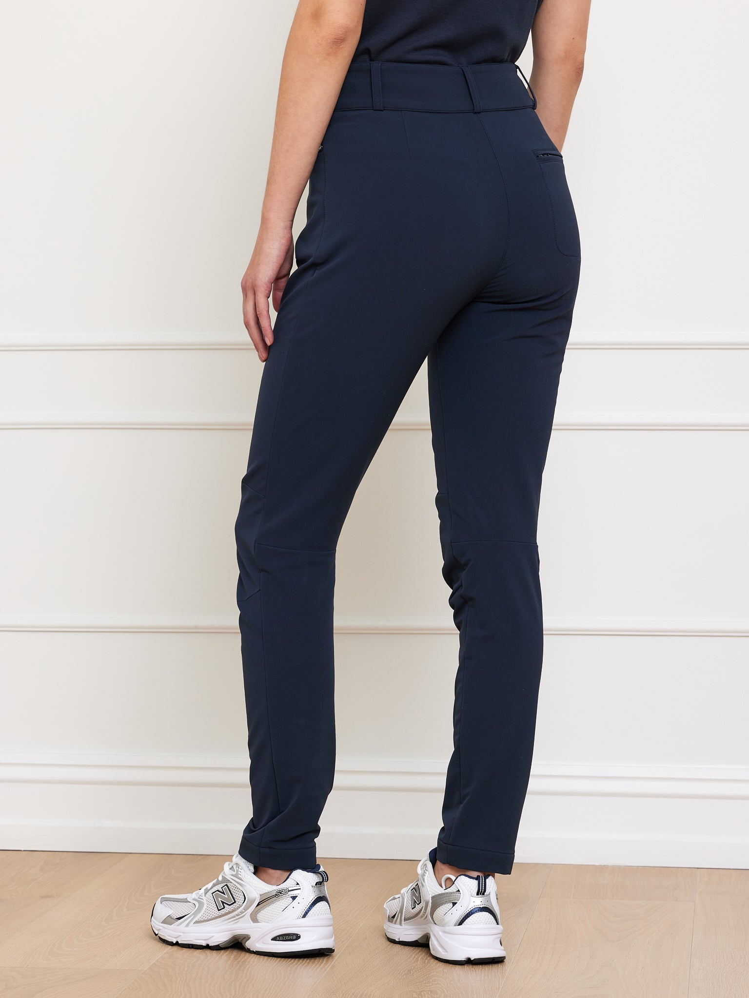 A person is wearing We Norwegians high-waisted navy Lista Pant Women, known for their sleek fit and innovative Schoeller fabric, paired with white sneakers against a white wall, one hand by their side.