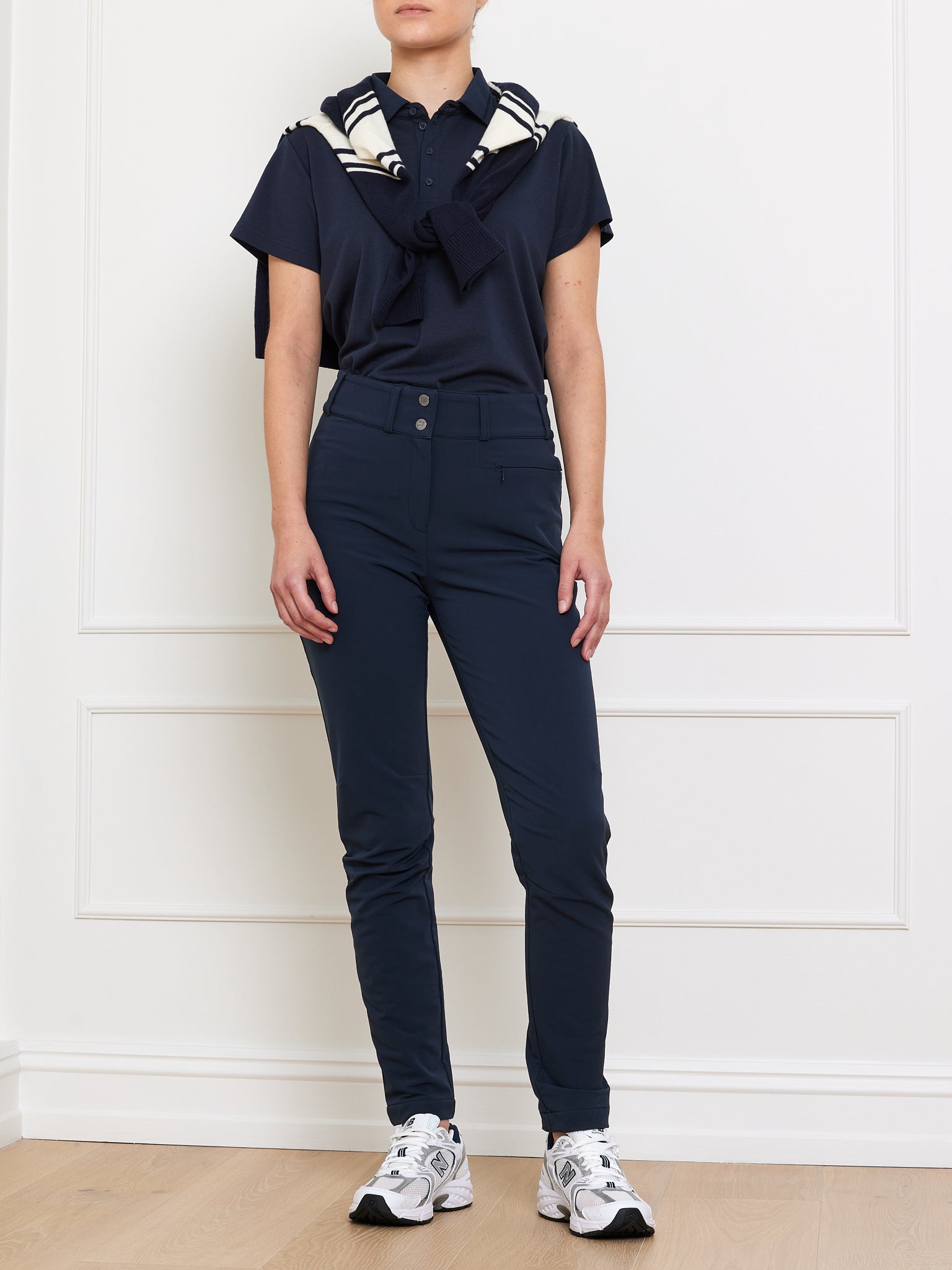 A person in a navy We Norwegians polo shirt and matching Lista Pant Women, with a striped sweater over their shoulders, wears white sneakers while standing against a white wall on light wood flooring.