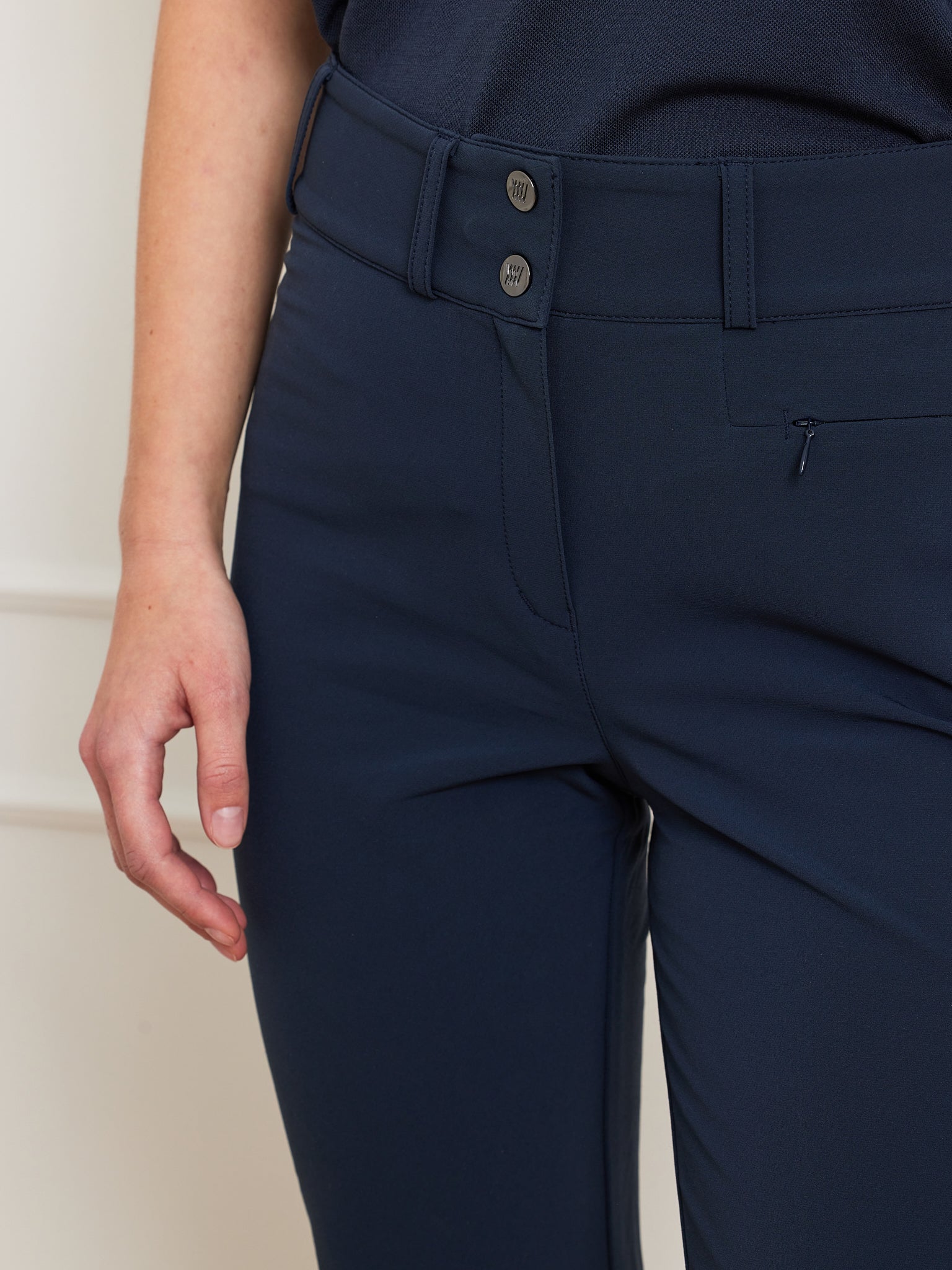Close-up of a person wearing dark navy, high-waist Lista Pant Women by We Norwegians, featuring a two-button waist enclosure. The top is partially visible; the left hand is relaxed at the side. The Schoeller fabric looks smooth and fitted.