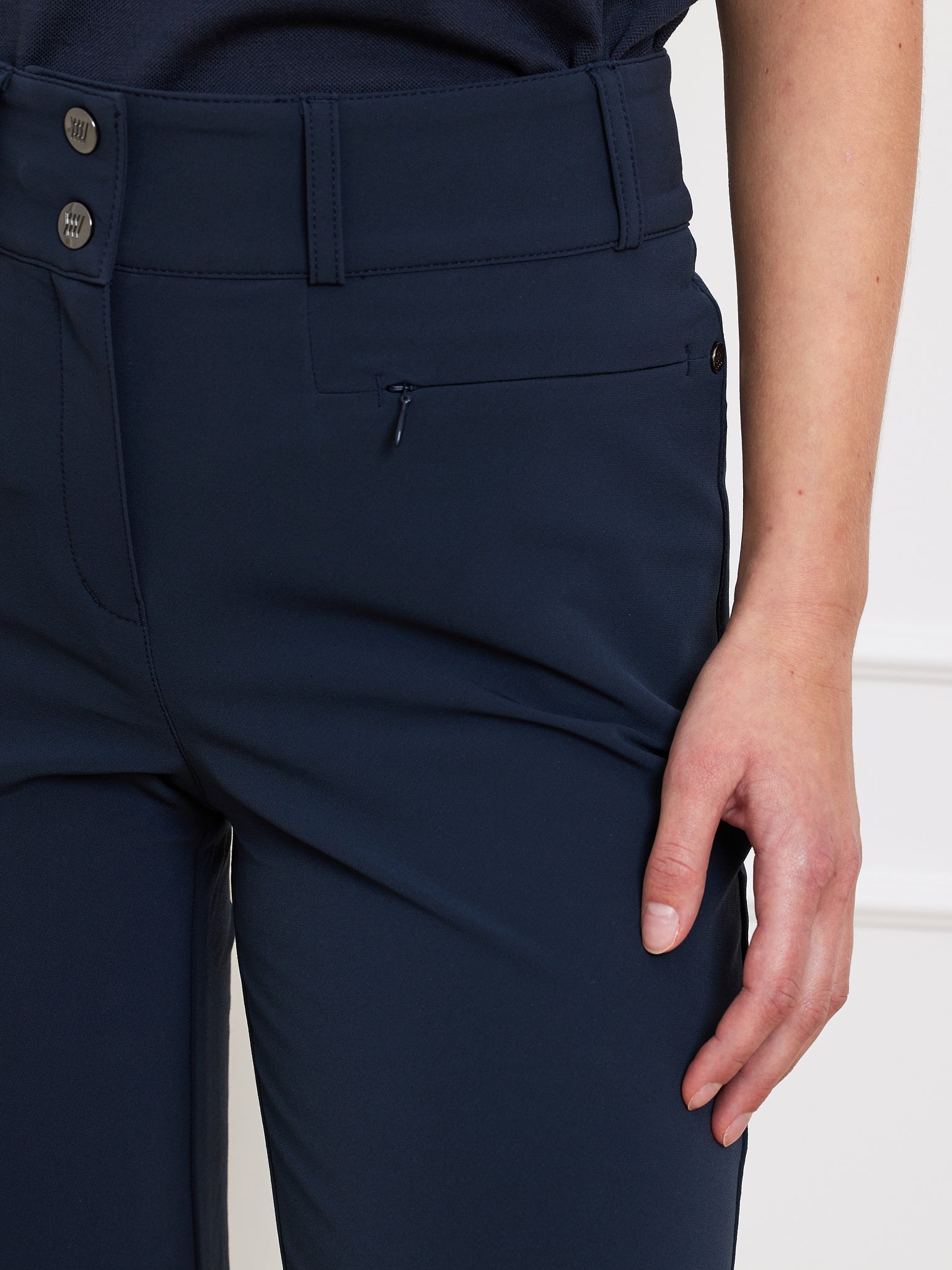 A person wears We Norwegians' Lista Pant Women in dark blue, made from high-waisted Schoeller fabric with a double-button closure and a small zippered pocket near the right hip. Their left hand rests beside the pocket.