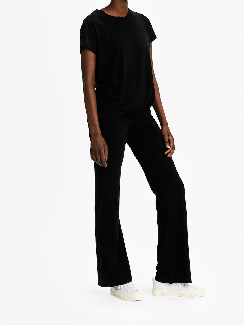A person wearing the Salt Classic Top Women Black from We Norwegians and black flared pants stands against a plain white background. They are also wearing white sneakers. Their arms are relaxed at their sides.