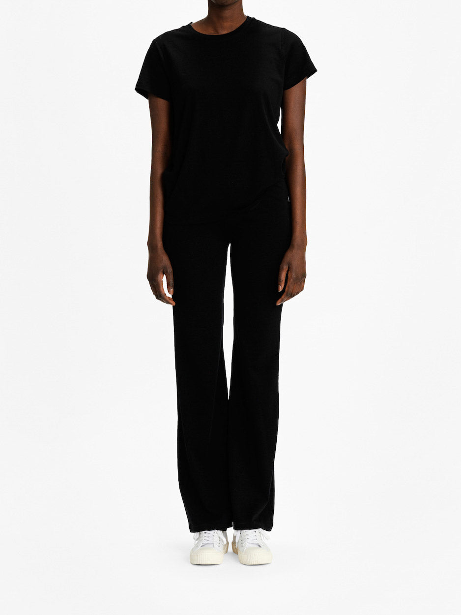 A person stands against a plain white background, wearing the Salt Classic Top Women Black by We Norwegians and black pants. Their arms are relaxed at their sides, and they are wearing white sneakers. Only their torso, arms, and legs are visible, not their face. This ensemble exudes Scandinavian Luxury Essentials style.
