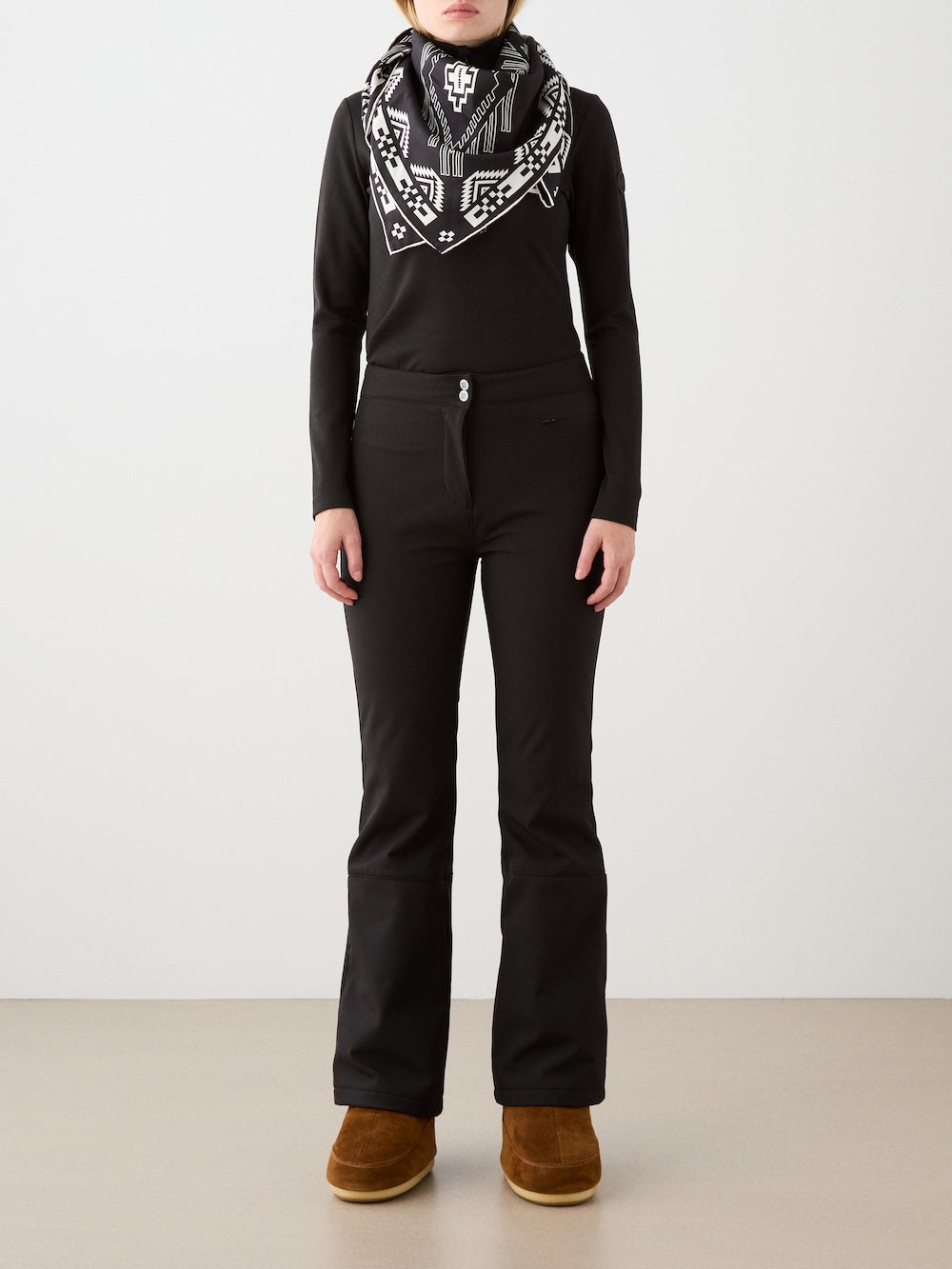 A person stands against a plain background wearing a black long-sleeve top, We Norwegians Tyin Bootleg Ski Pant Women, a black and white patterned scarf, and brown boots. Their hands rest at their sides.
