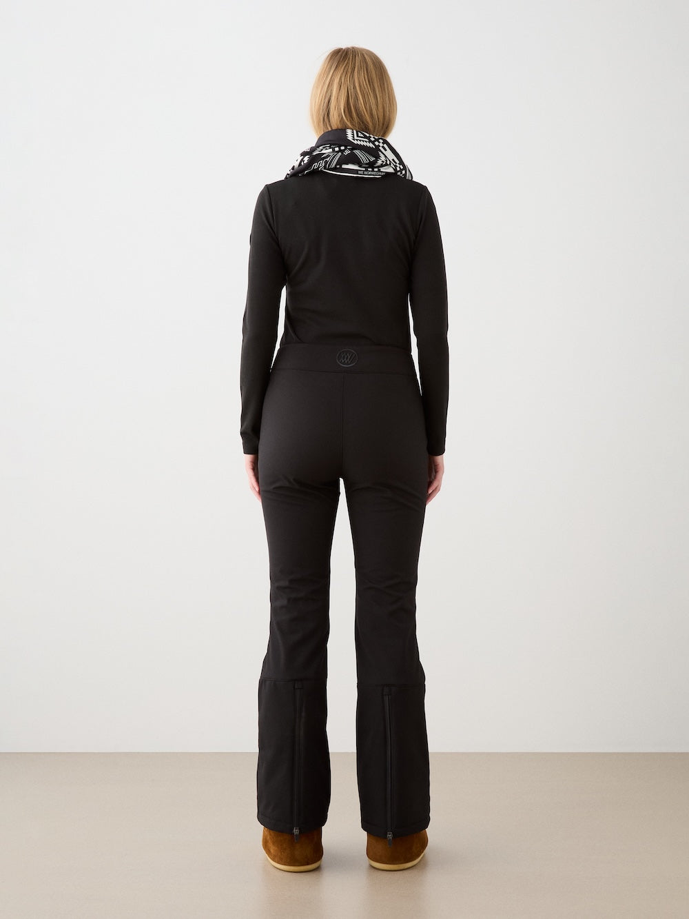 A woman with blonde hair faces away, wearing a black long-sleeve top, We Norwegians Tyin Bootleg Ski Pant Women, a black-and-white patterned scarf, and brown boots against a plain background.