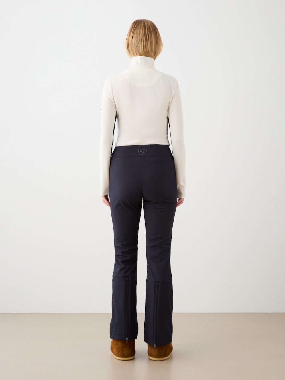 A woman with shoulder-length blonde hair, in a cream turtleneck and We Norwegians Tyin Bootleg Ski Pant for women, stands facing a white wall on a light brown floor, paired with brown boots.
