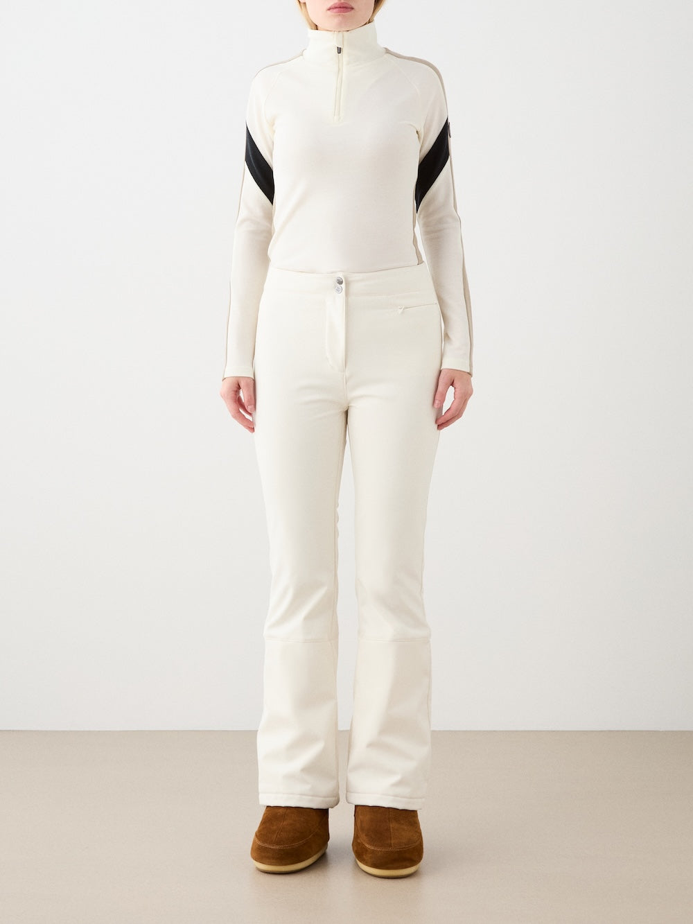 A person stands facing forward against a plain background, wearing We Norwegians Tyin Bootleg Ski Pant Women, a fitted cream long-sleeve top with black diagonal sleeve stripes, and brown boots. The person's head is cropped out of the image.
