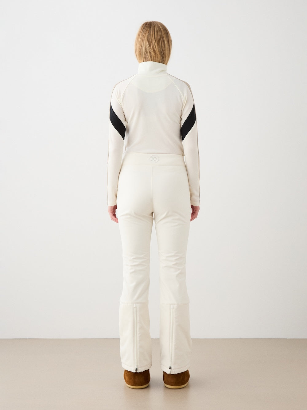 A woman with light brown hair faces away, wearing a fitted white ski outfit featuring We Norwegians’ Tyin Bootleg Ski Pant Women and a water-repellent soft shell with brown boots, set against a plain white background.