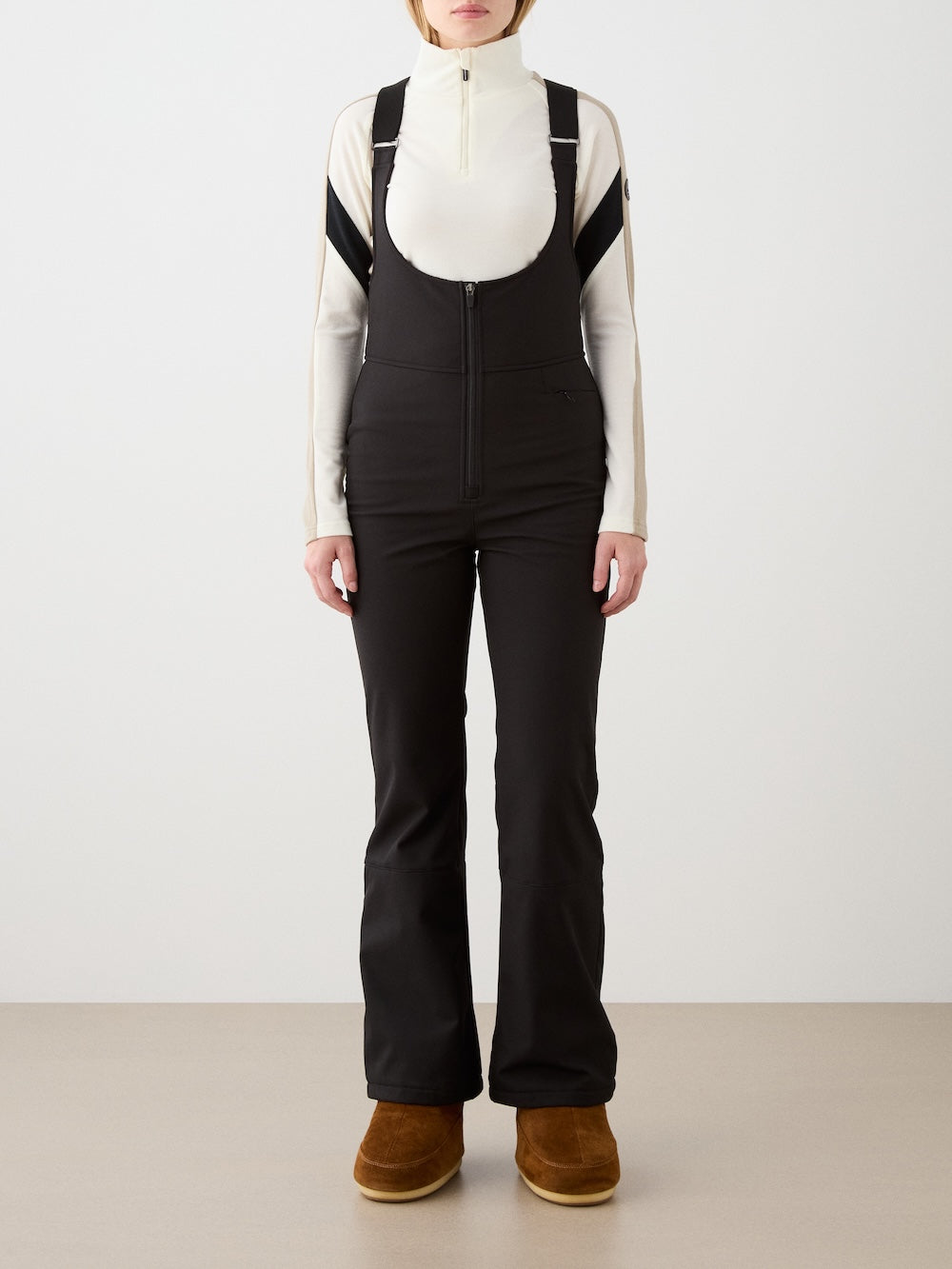 A person faces forward against a plain background, wearing We Norwegians Tyin Bib Pant Women—black high-waisted fitted bib ski pants with a front zipper—paired with a cream long-sleeve zip-up turtleneck and brown suede boots.
