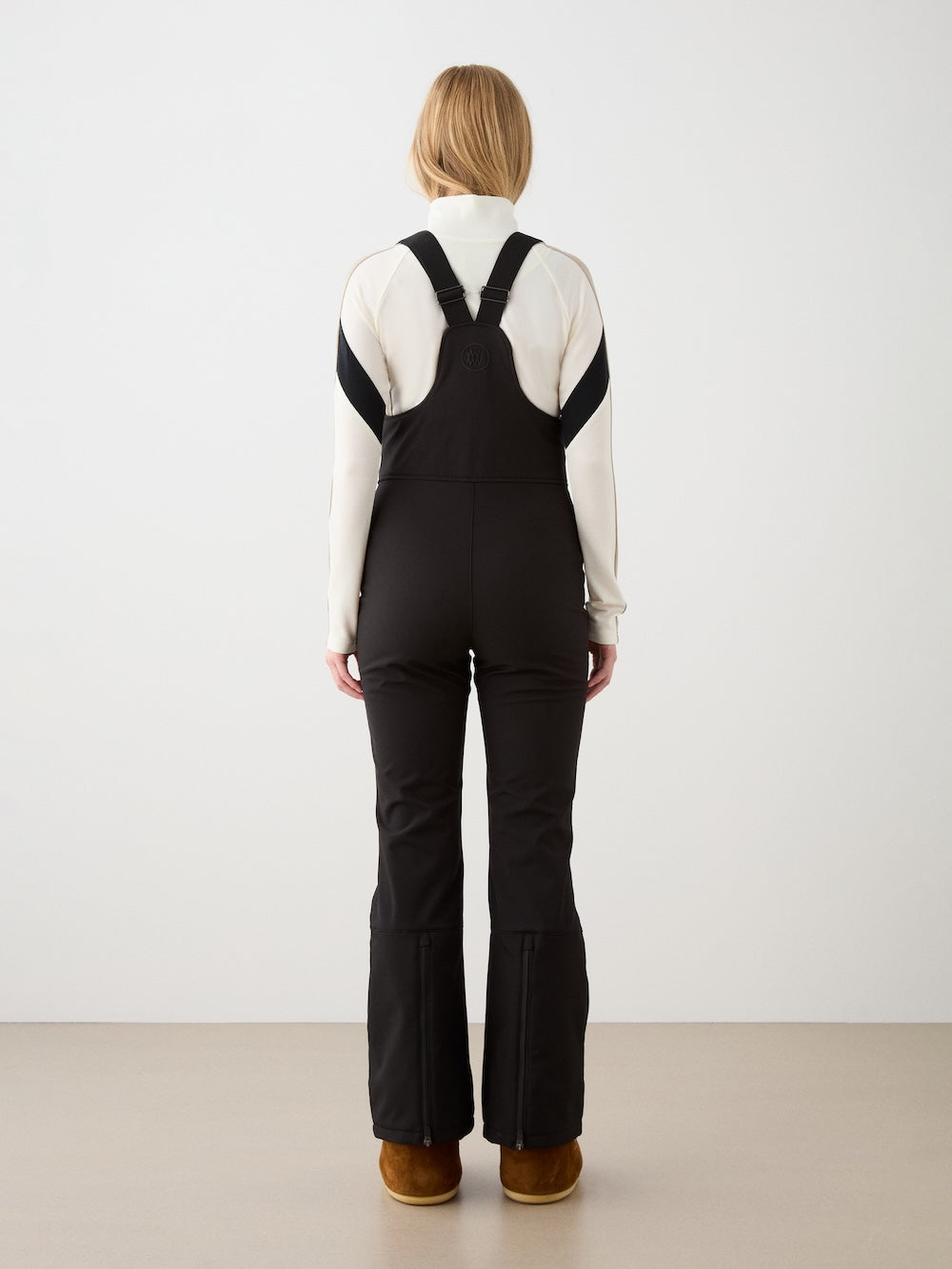 A person with blond hair, wearing a white long-sleeve top and We Norwegians Tyin Bib Pant Women in black with cross-back straps, plus brown boots, stands facing a plain white wall with their back to the camera.