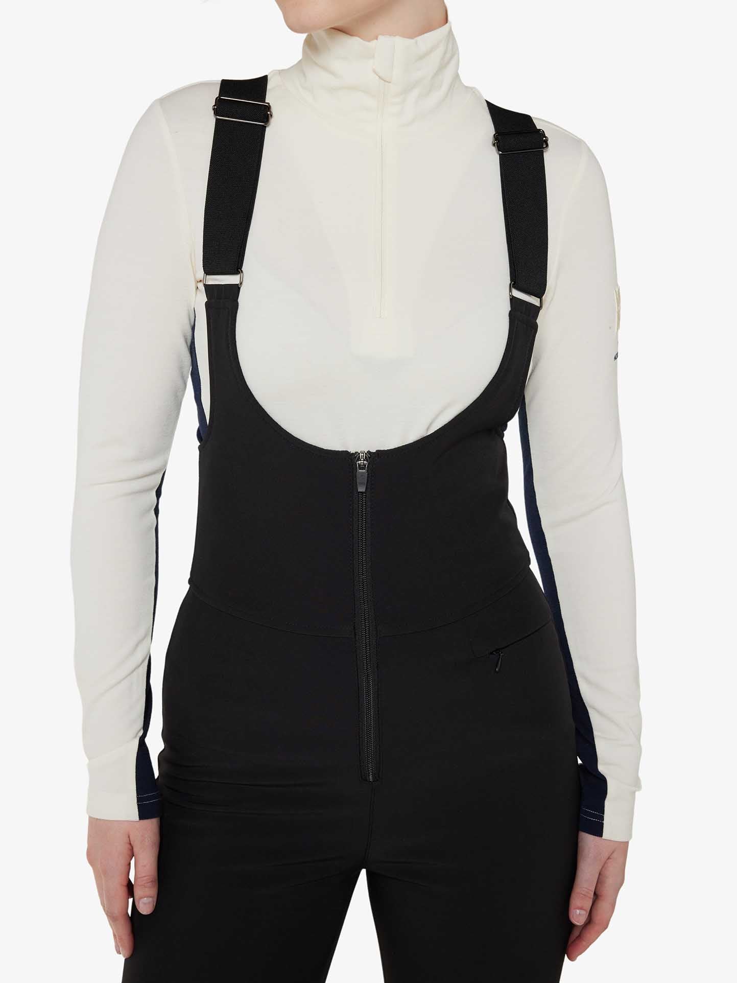 A person is shown from the shoulders to the hips, wearing a white turtleneck long-sleeve shirt and Tyin Bib Pant Women in black. The high-waisted ski pants, from We Norwegians, are made of water repellent soft shell fabric and feature adjustable straps over the shoulders and a front zipper. The background is plain white.