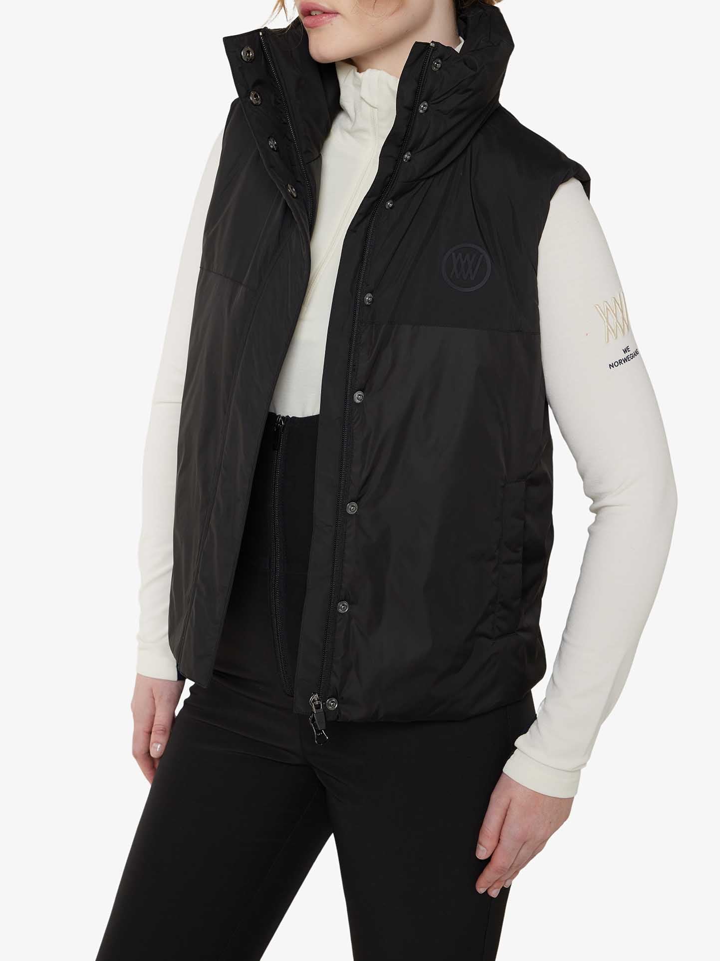 A person wearing a black quilted vest over a white long-sleeve shirt with black pants. The vest has a high collar and is fastened with buttons. The individual is styled in the Tyin Bib Pant Women by We Norwegians, which enhances the high-waisted shape and is crafted from water-repellent soft shell fabric. The shirt features a logo on the left sleeve, while the person's face is not fully visible.