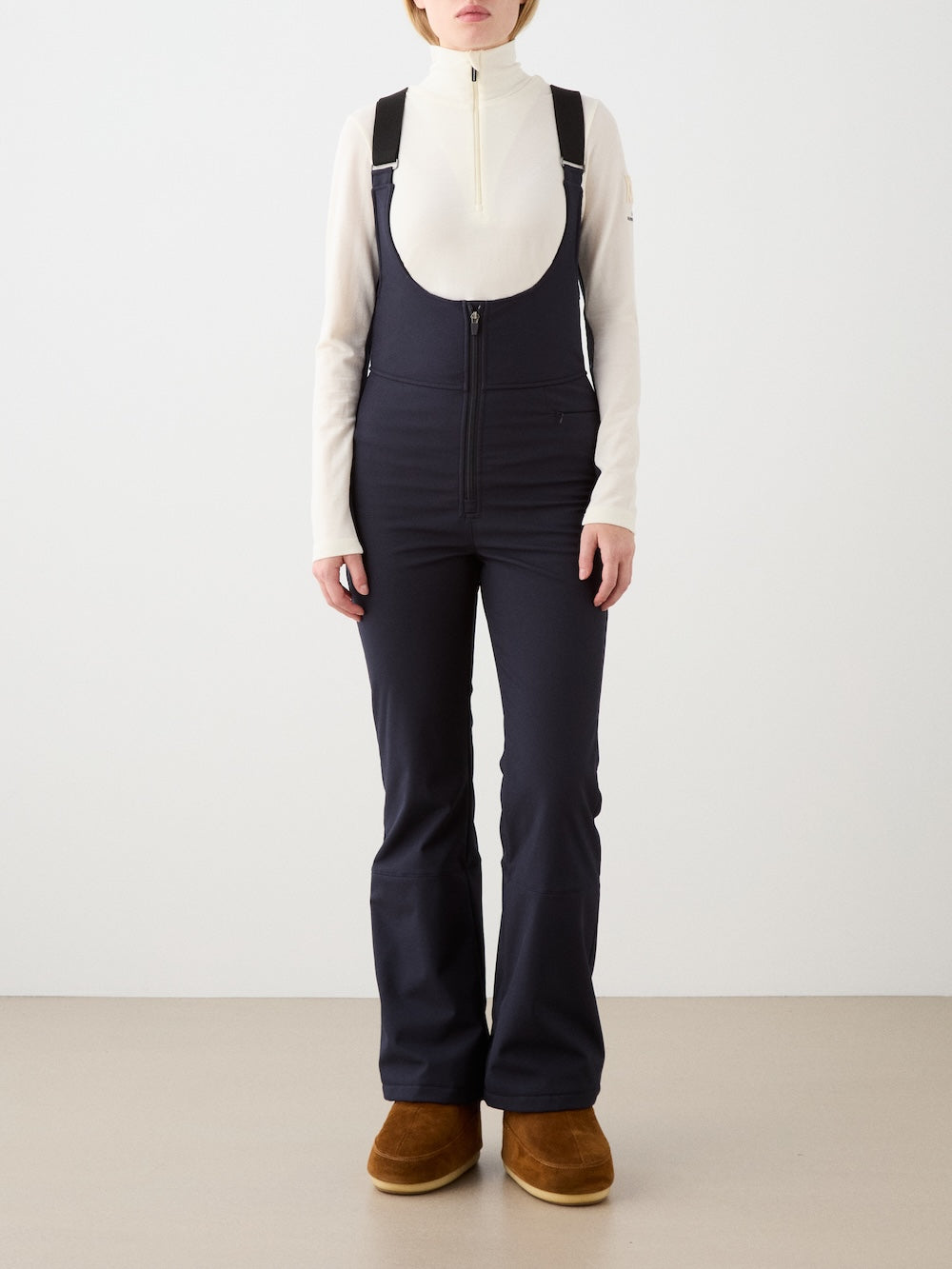 A woman stands indoors in We Norwegians Tyin Bib Pant Women—navy, high-waisted, water-repellent bib pants with black straps. She pairs them with a cream zip-neck top and brown fur-lined boots against a plain background.