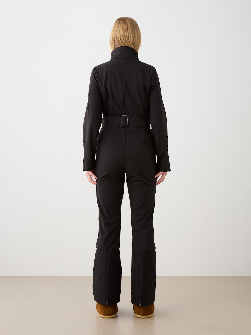 A person with straight, shoulder-length hair faces away against a light background, wearing the fitted black Tyin Ski Suit Women by We Norwegians in a water-repellent soft shell and brown shoes.