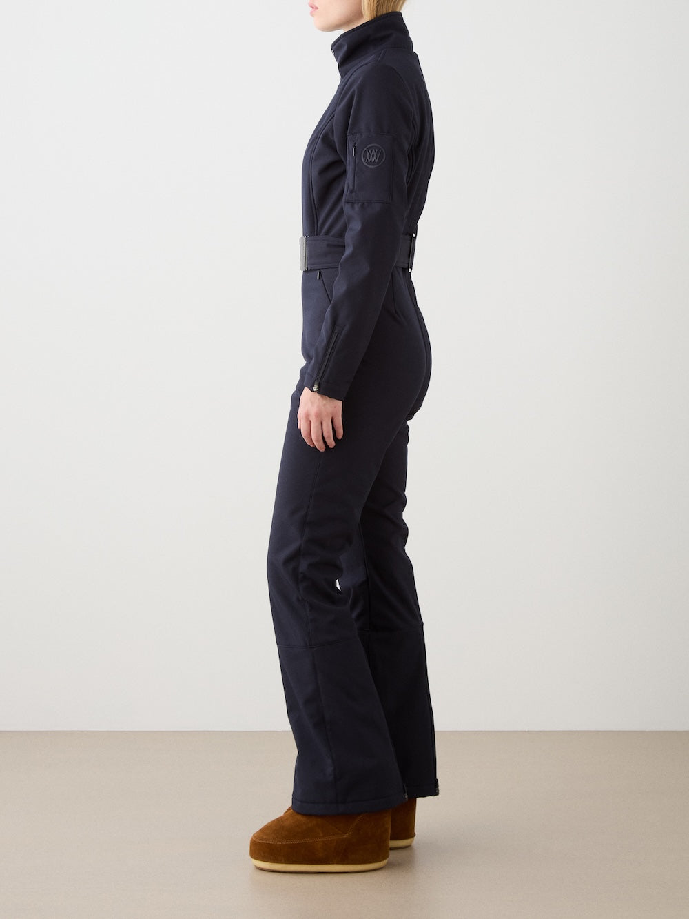 A person stands sideways wearing the fitted We Norwegians Tyin Ski Suit Women in dark navy—a water-repellent soft shell with a belt and brown boots—against a plain white wall and light floor.
