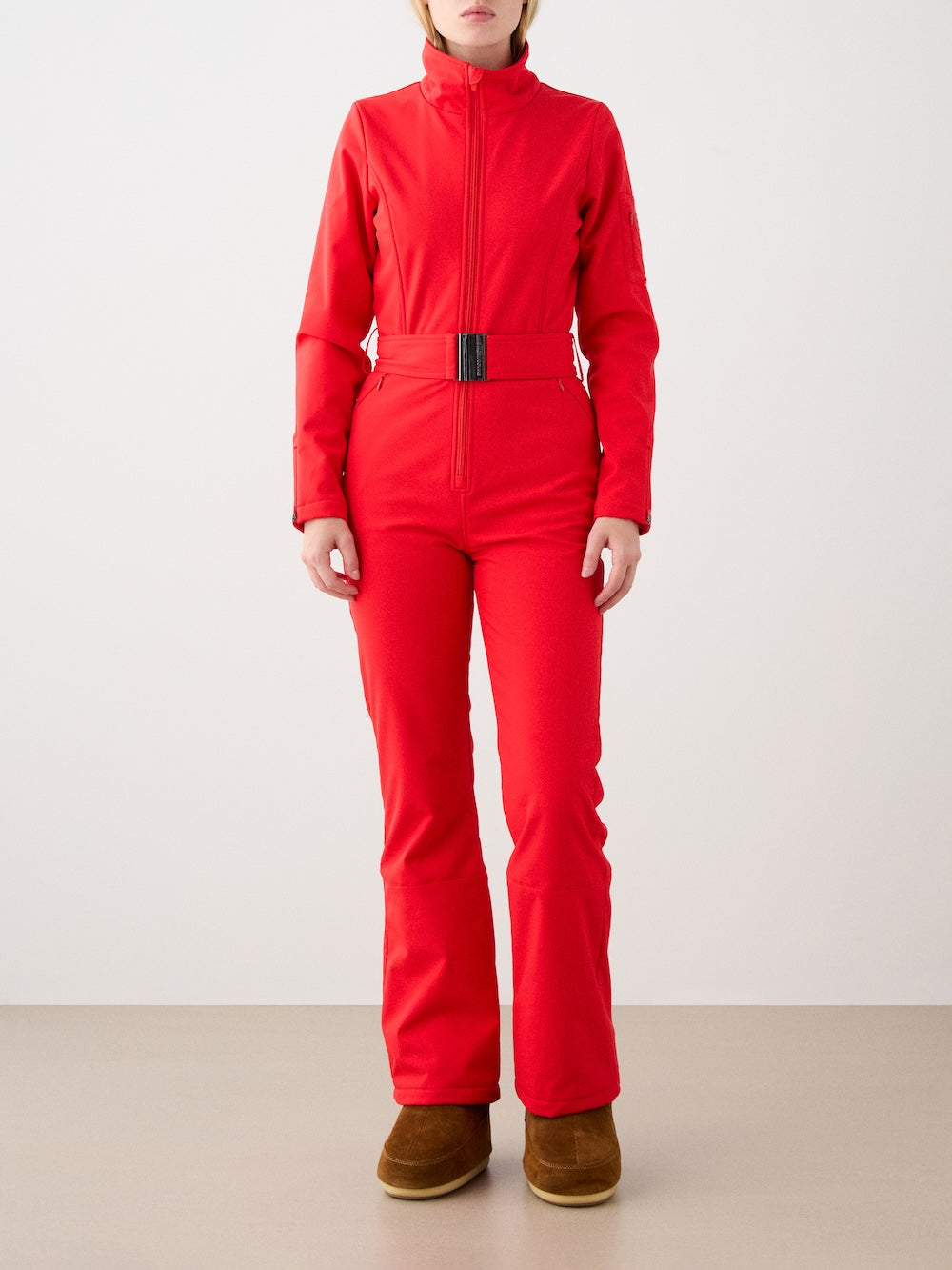 A person stands against a plain background in a bright red We Norwegians Tyin Ski Suit Women with a high collar and belted waist, made from water-repellent soft shell, paired with brown flat winter boots. Their face is partially out of frame.