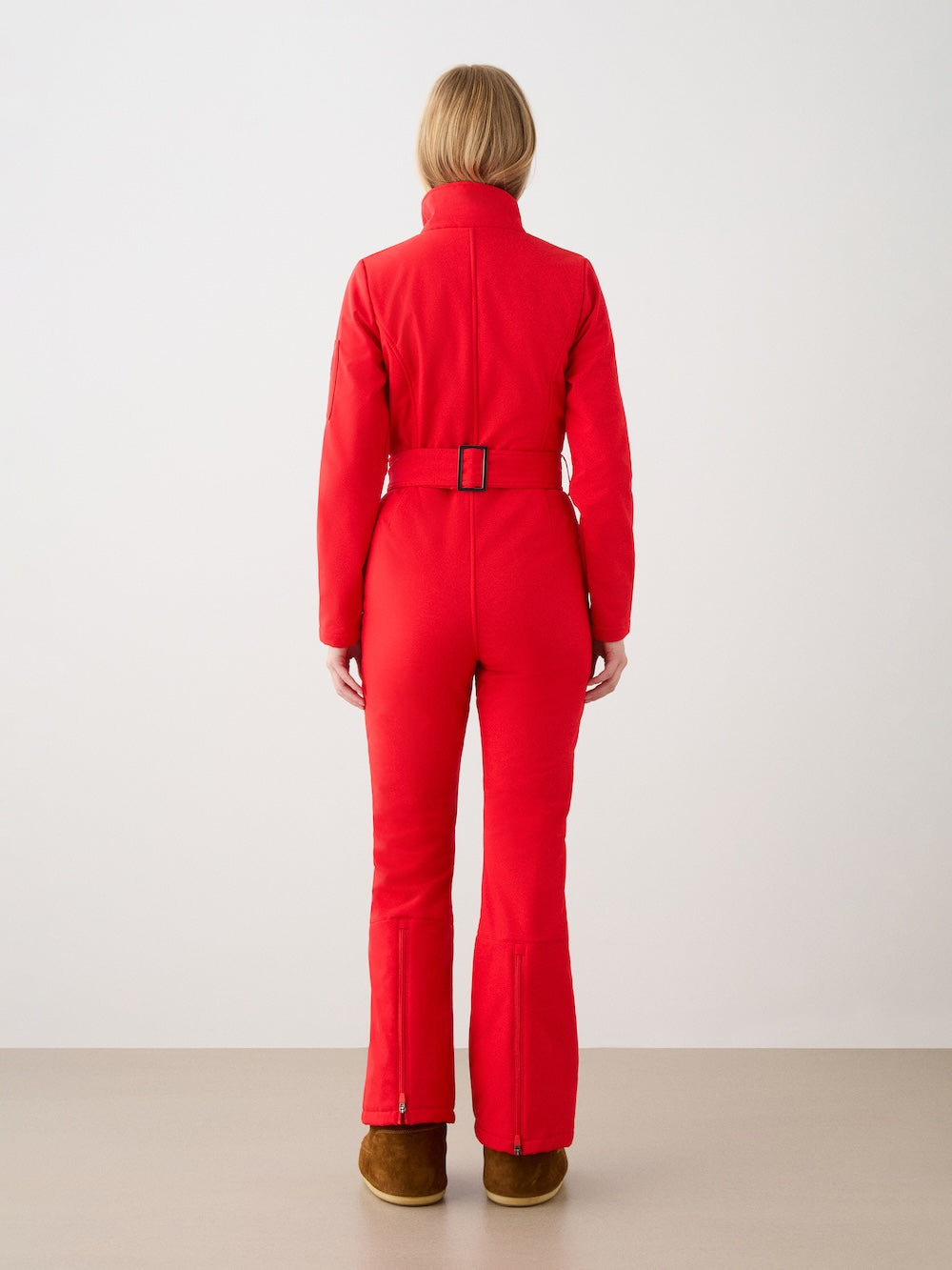 A woman with short blonde hair faces away, wearing a fitted red We Norwegians Tyin Ski Suit made from water-repellent soft shell fabric and brown boots, set against a plain light background.