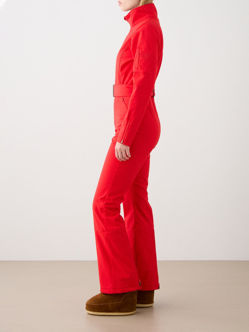 A person stands sideways in the fitted, bright red Tyin Ski Suit Women by We Norwegians, featuring a high collar and belt. Made from water-repellent soft shell for style and function, it’s paired with brown ankle boots against a plain light background.