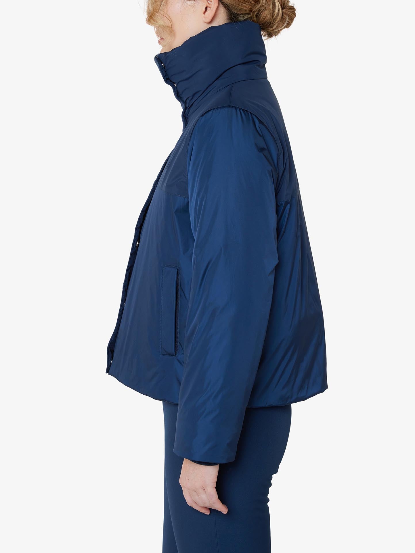 A person with light-colored hair styled in a bun is shown in a profile view against a white background, wearing the We Norwegians Filefjell Ski Jacket Women and matching blue pants. The high collar of the navy blue, water-repellent jacket is zipped up.