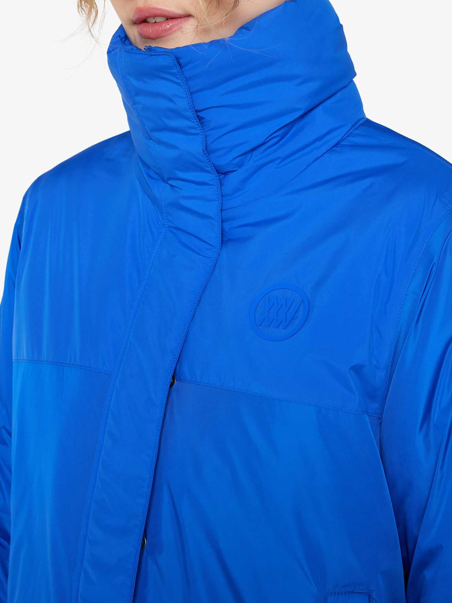 A close-up view of a person wearing a We Norwegians Filefjell Ski Jacket Women in bright blue, featuring a high collar. The person has light, wavy hair partially visible at the top. An embossed "XXV" logo is on the chest of the jacket, which is made from water-repellent fabric to keep you dry. The background is white.