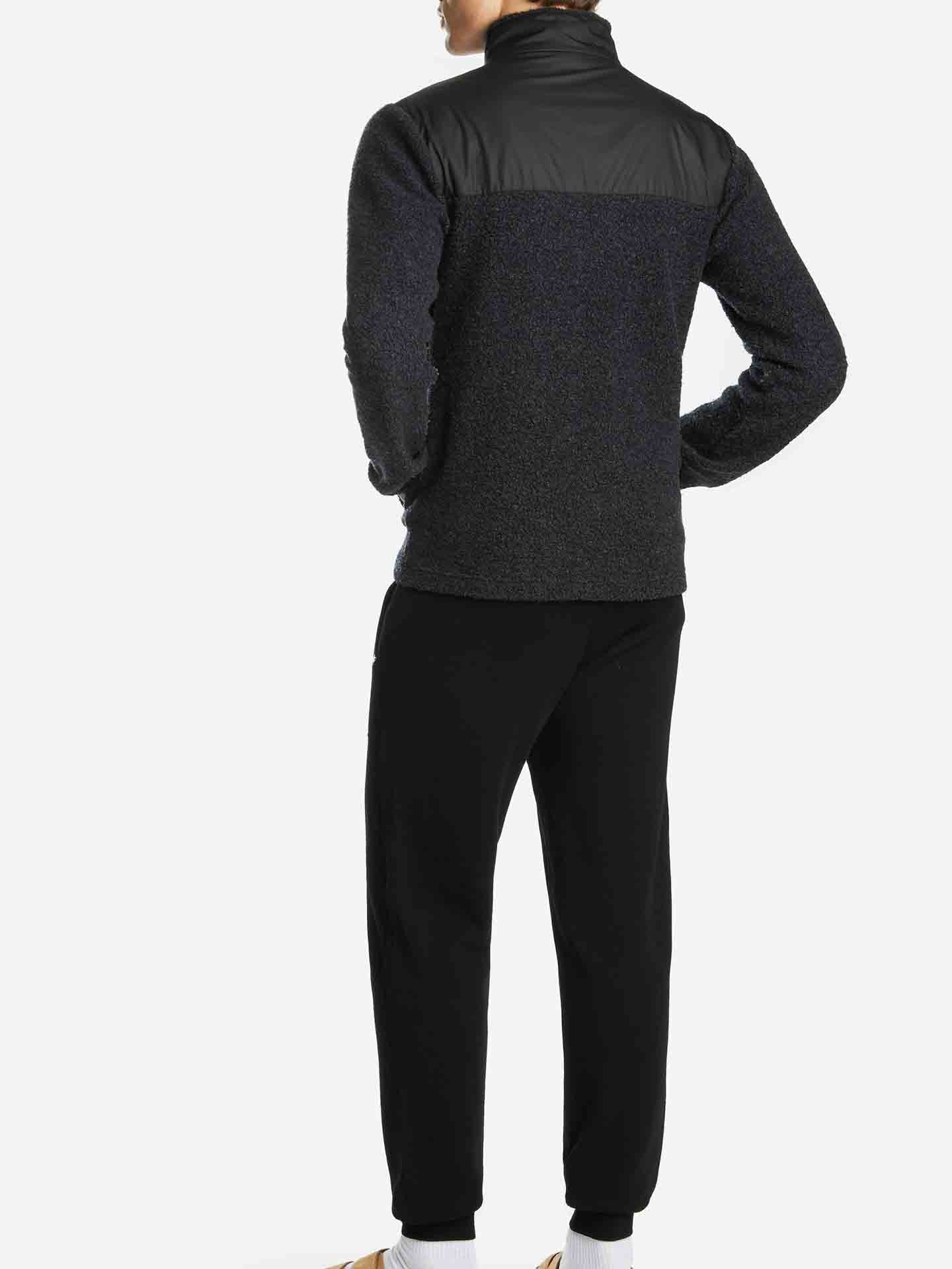 A person is standing with their back to the camera, wearing a dark gray "Alta Jacket Men" fleece from We Norwegians, featuring black shoulder panels, paired with black pants. The background is plain white.