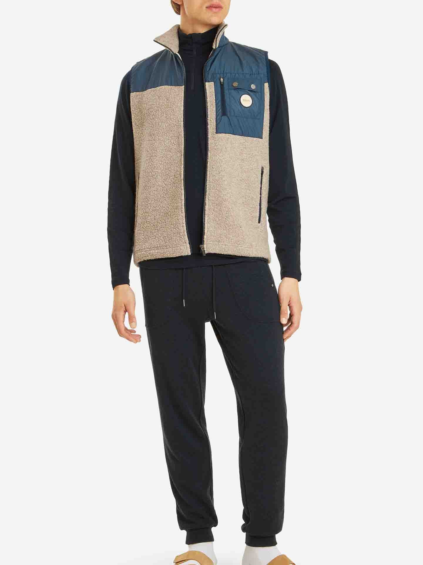 A person is wearing the We Norwegians Alta Vest Men in beige and navy blue over a black long-sleeve shirt and black jogger pants. The individual is also sporting beige slip-on shoes. Their hands are relaxed by their sides, and the background is plain white.