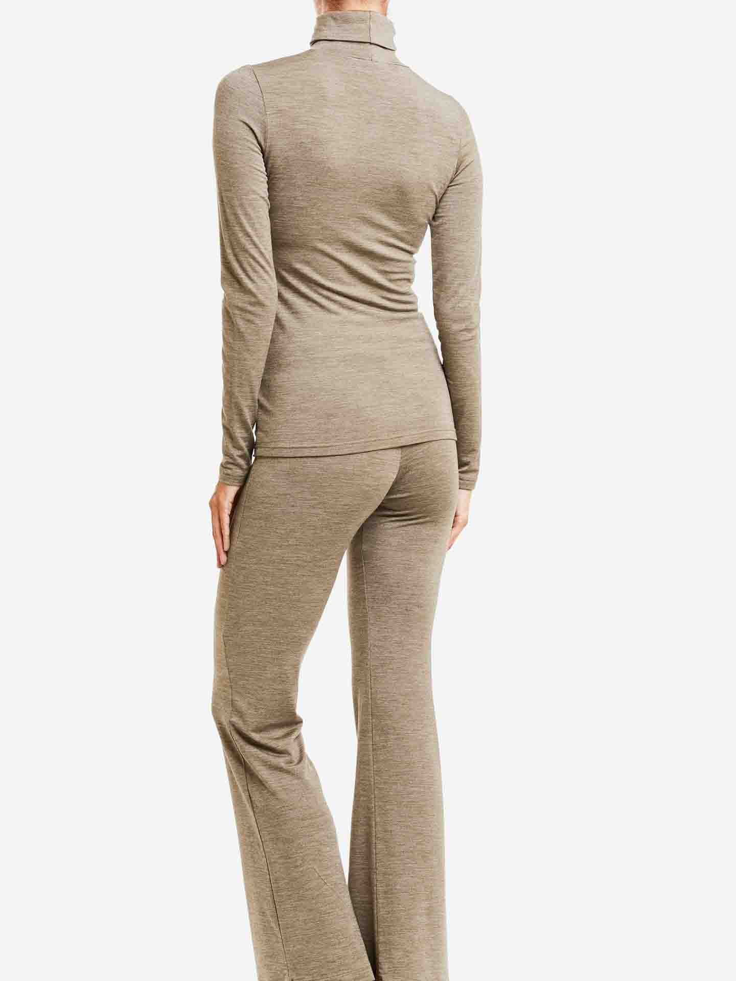 A person is standing facing away from the camera, wearing a light brown, long-sleeved Hygge Turtleneck Women base layer and matching pants from We Norwegians. The slightly flared, form-fitting outfit appears comfortable and is made from cozy merino wool.