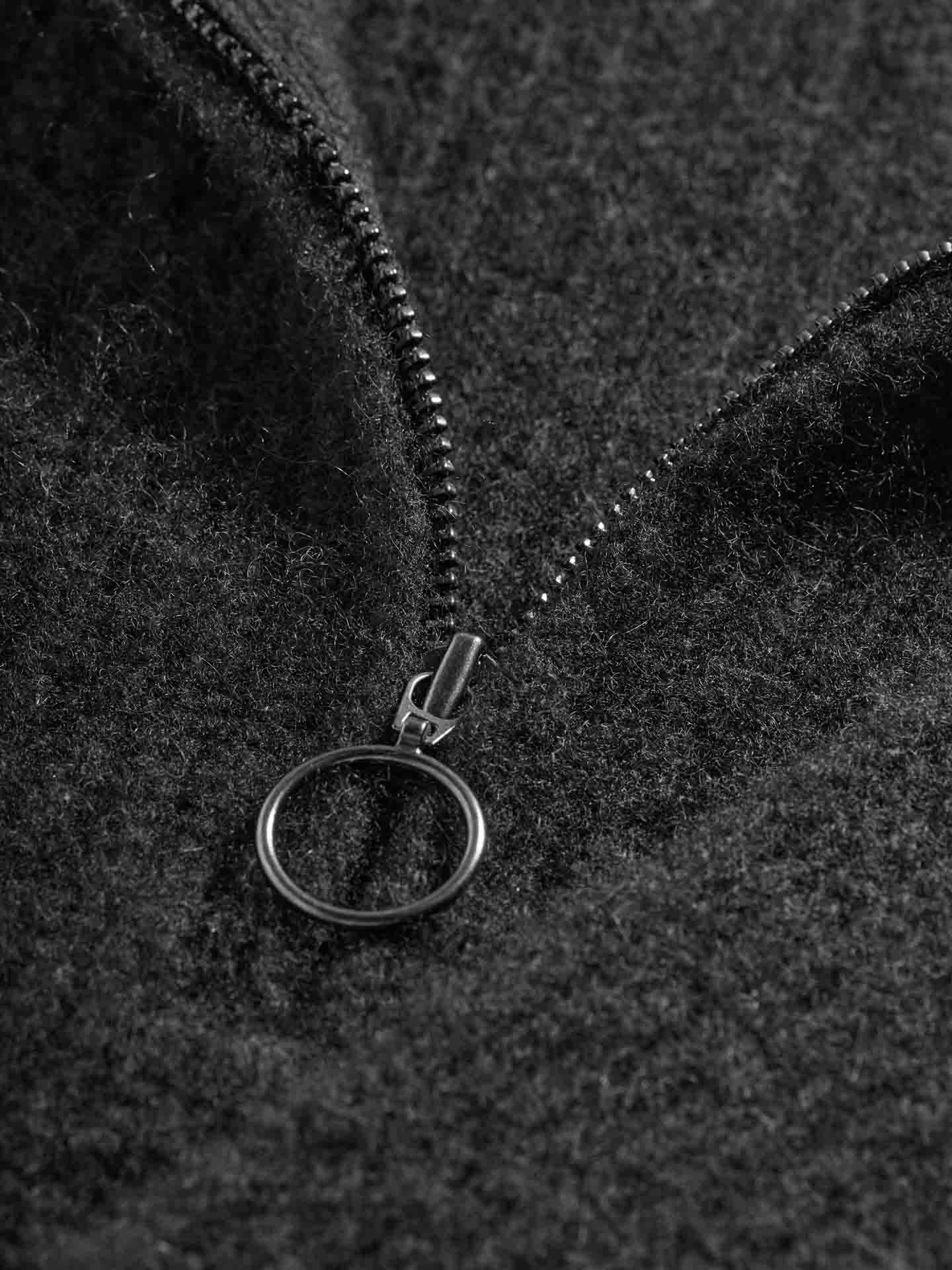 Close-up image of the We Norwegians Blefjell Cashmere Bib Women in a black metal half-zip with a circular pull tab against dark grey fabric. The soft, fuzzy texture of the cashmere bib contrasts with the shiny metal zipper.