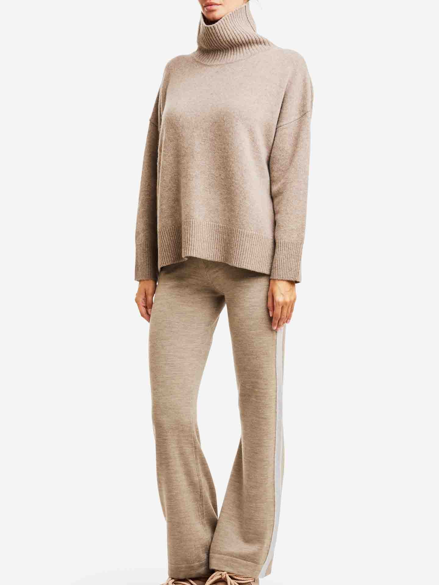 Dressed in the Blefjell Cashmere Sweater by We Norwegians, paired with matching wide-leg pants with white side stripes, a person exudes timeless style. The cozy beige outfit appears luxuriously soft against a plain white background.