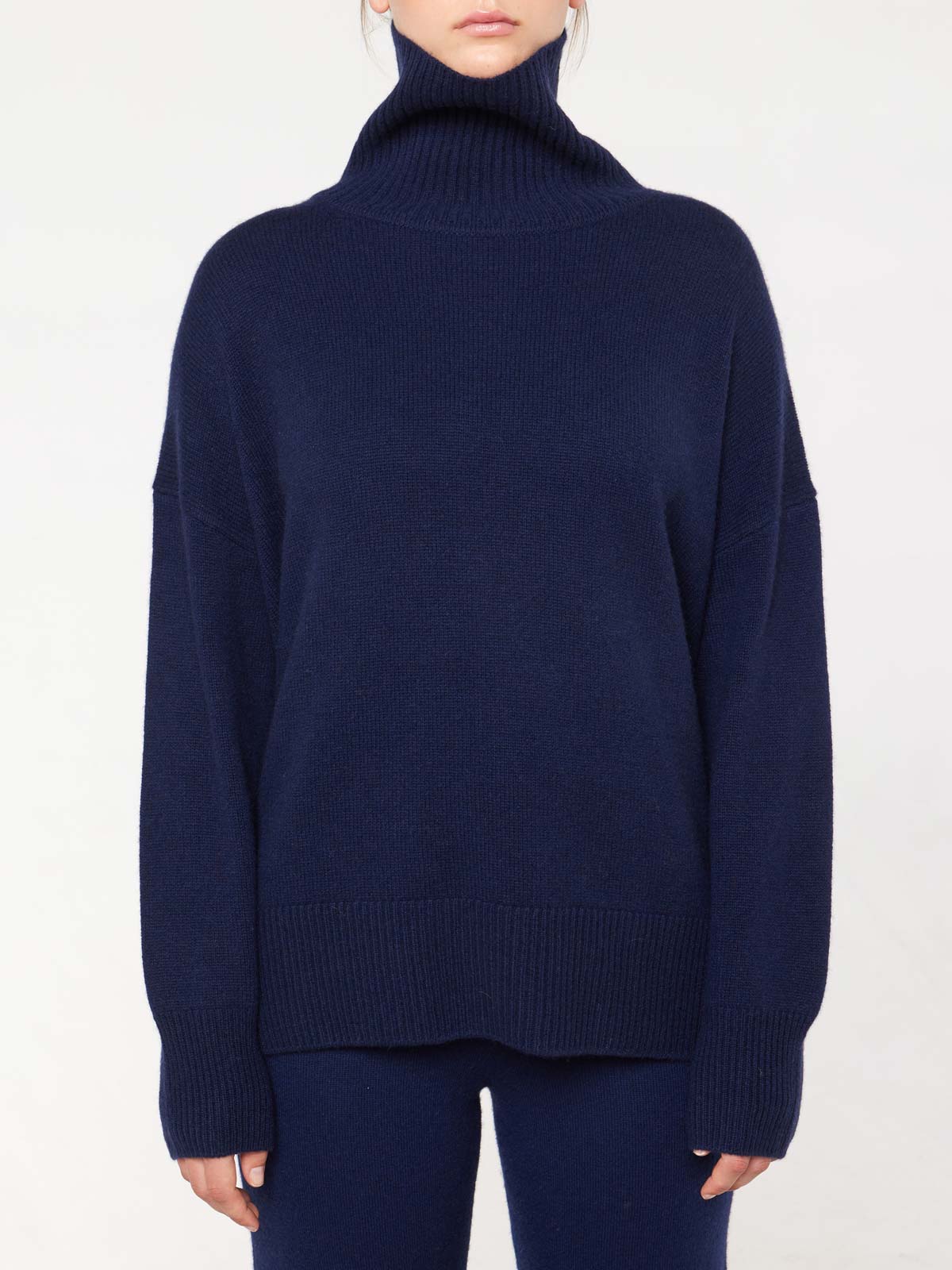 A person in a We Norwegians dark blue Blefjell Cashmere Sweater with long sleeves and matching pants stands against a plain white background, exuding timeless style.