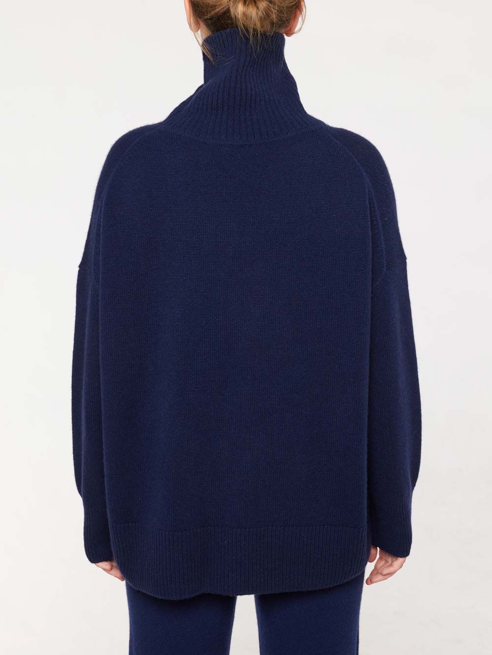 A person with their hair up is seen from the back wearing We Norwegians' Blefjell Cashmere Sweater for women in dark blue, oversized and styled with a turtleneck, standing against a plain white background, embodying timeless style.
