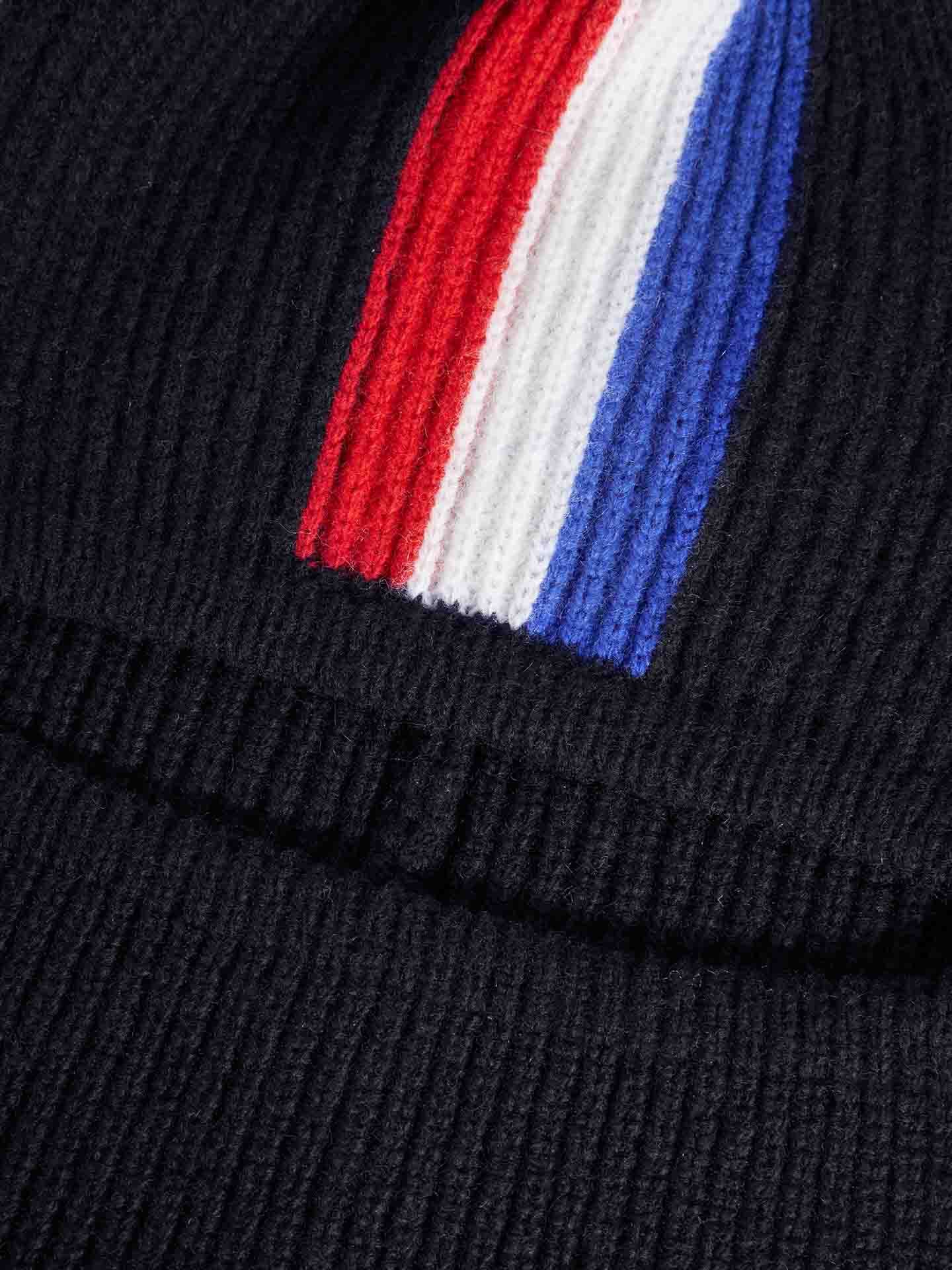 Close-up of the Apres Ski Balaclava by We Norwegians, showcasing a black knit fabric with a centered vertical emblem featuring red, white, and blue stripes. Evoking 70s ski fashion, the stripes run from top to bottom within the frame. The prominent texture of the knitted material conjures thoughts of après-ski wear.