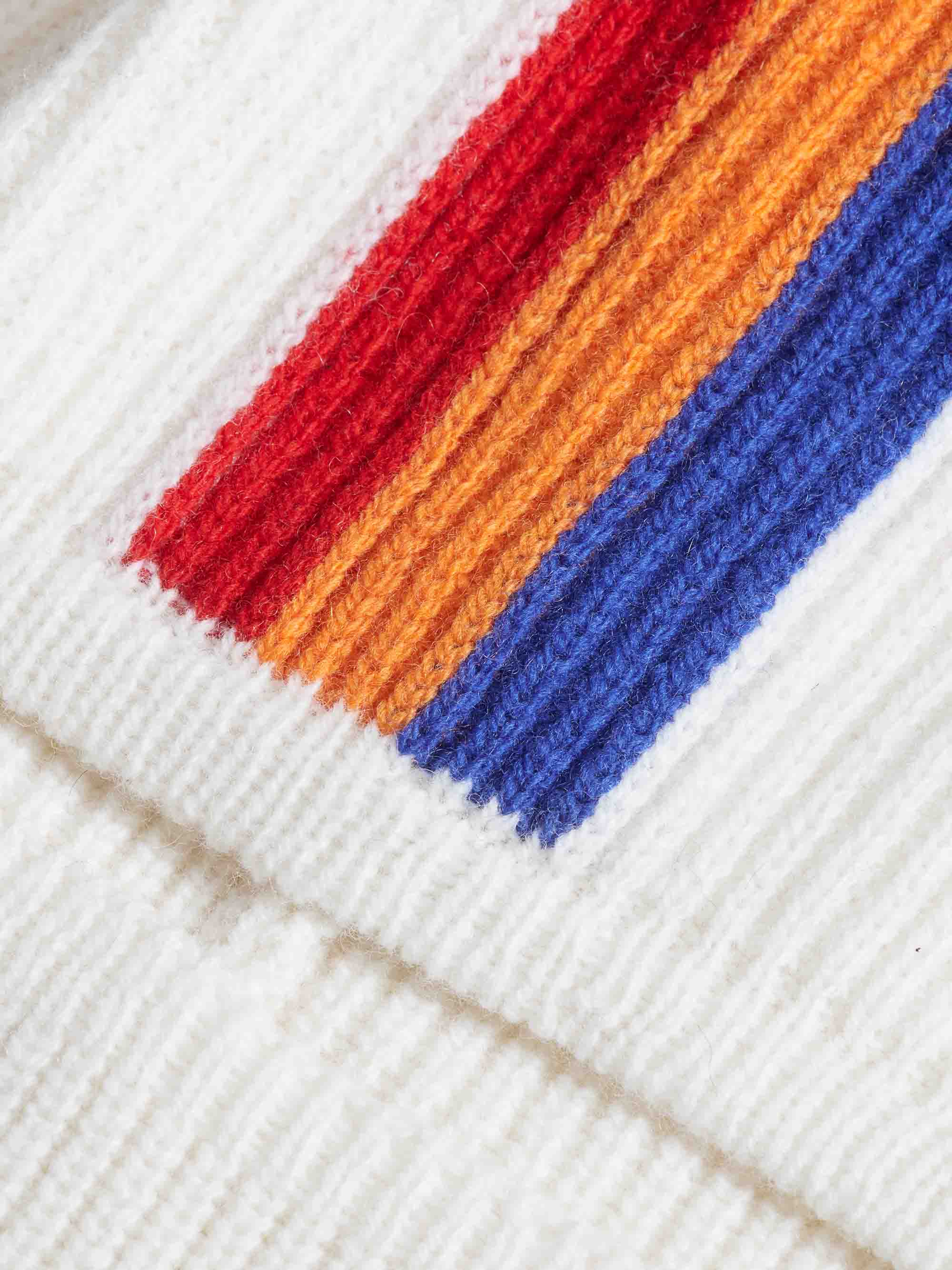 Close-up image of the Apres Ski Balaclava by We Norwegians, showcasing its knitted fabric with thick, vertical stripes in red, orange, and blue on a white background. The textured merino wool and vivid colors create a visually striking pattern reminiscent of 70s ski fashion.