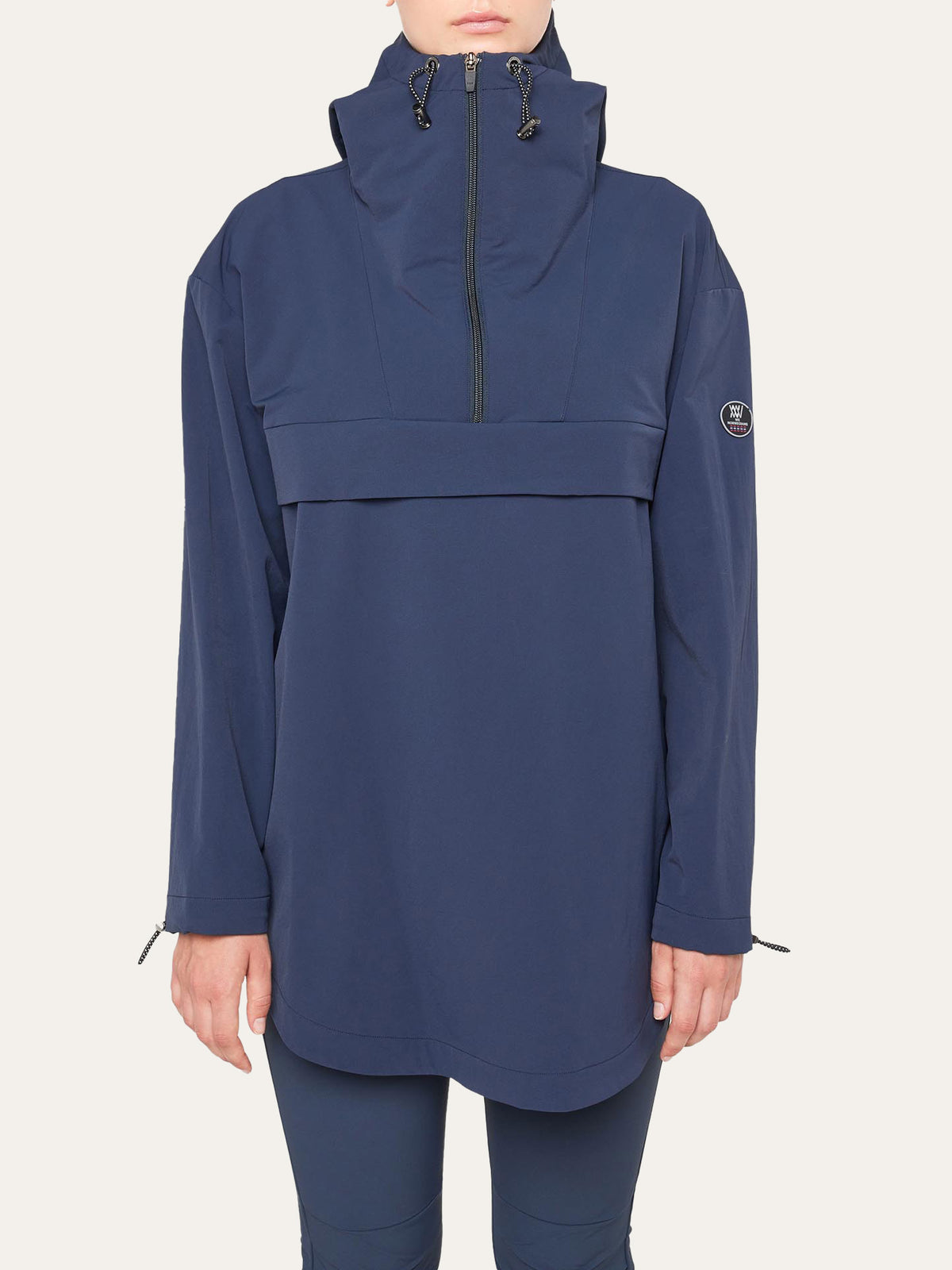 A woman wears the We Norwegians Beito Anorak— a loose-fitting, long-sleeve, navy jacket with a high zip collar and drawstrings, inspired by Norwegian outdoor style. A patch accents one sleeve. She stands against a plain light background.