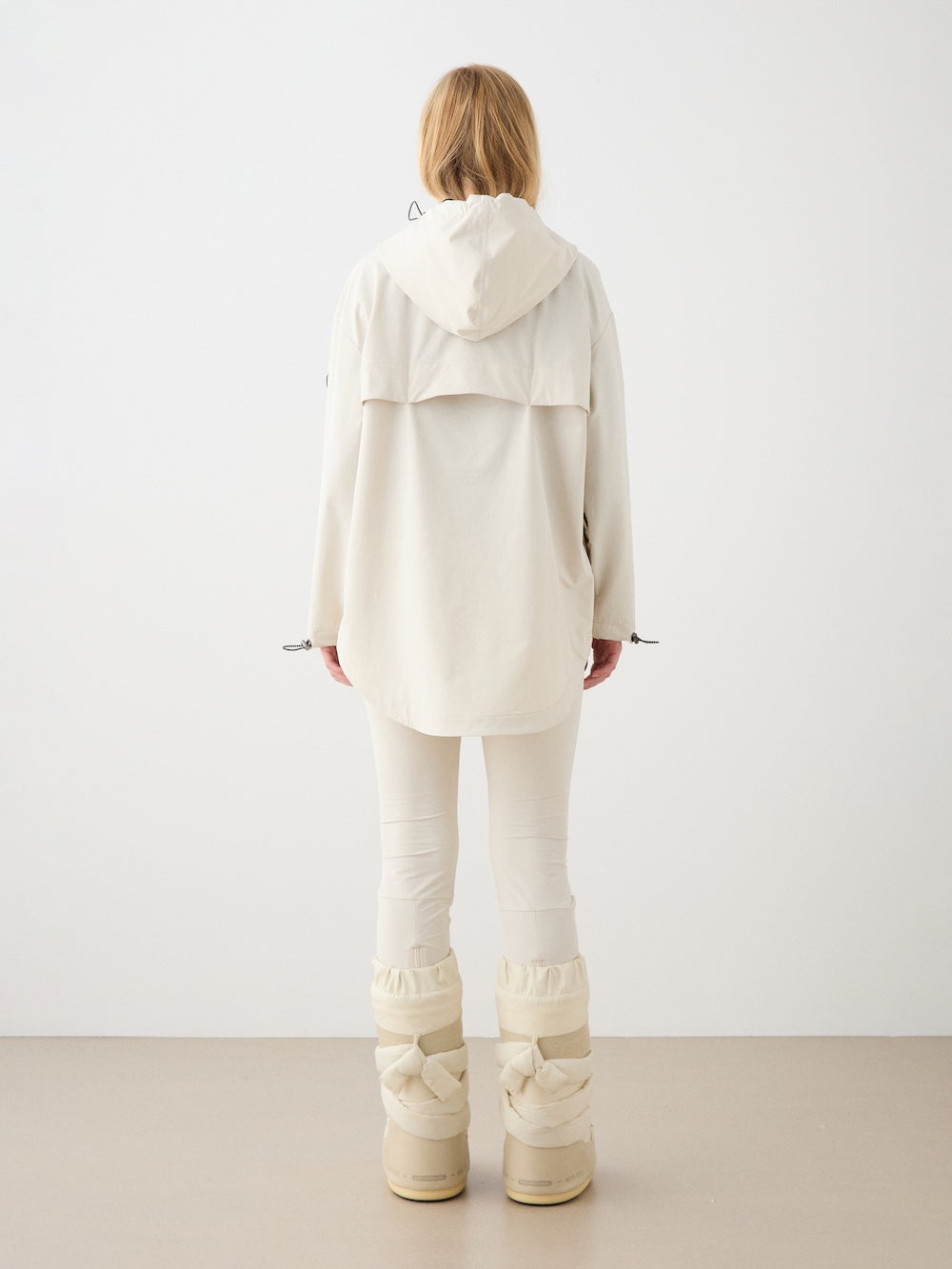 A person with long blonde hair faces away, wearing the We Norwegians Beito Anorak for women with matching pants and beige boots against a plain background, embodying Norwegian outdoor style.