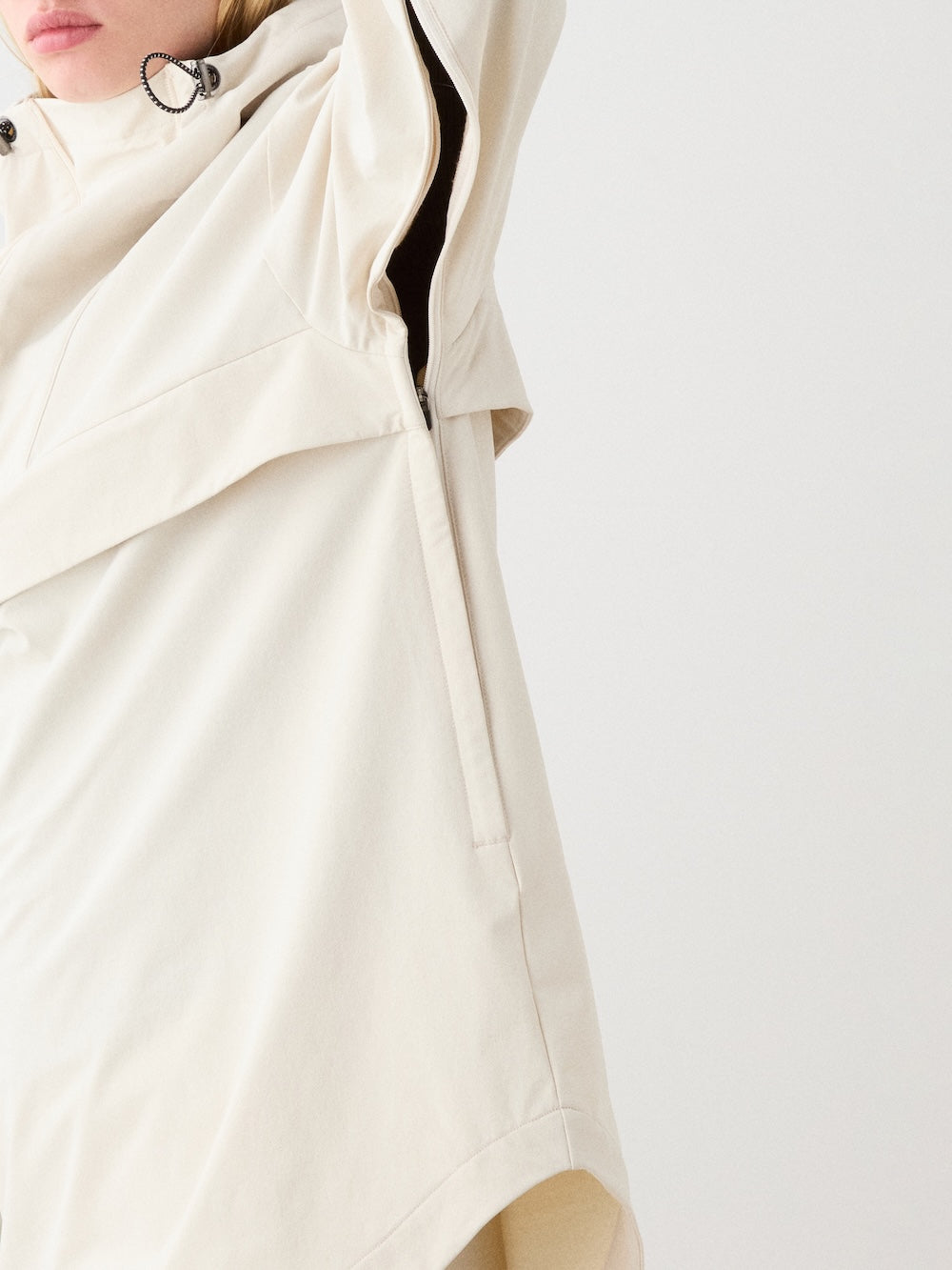 A close-up of a person raising one arm, partially obscuring their face, wearing the We Norwegians Beito Anorak Women— a loose, light beige ski anorak with high collar and black drawstring—against a plain white background.