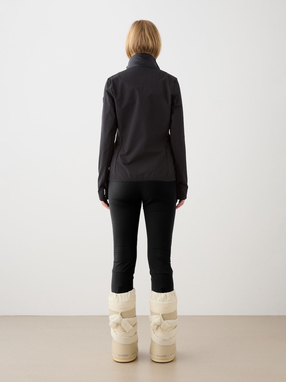 A woman with short light brown hair wears the We Norwegians Beito Jacket Women in black, black pants, and tall off-white boots, standing with her back to the camera in a minimal indoor space with a beige floor and white wall.