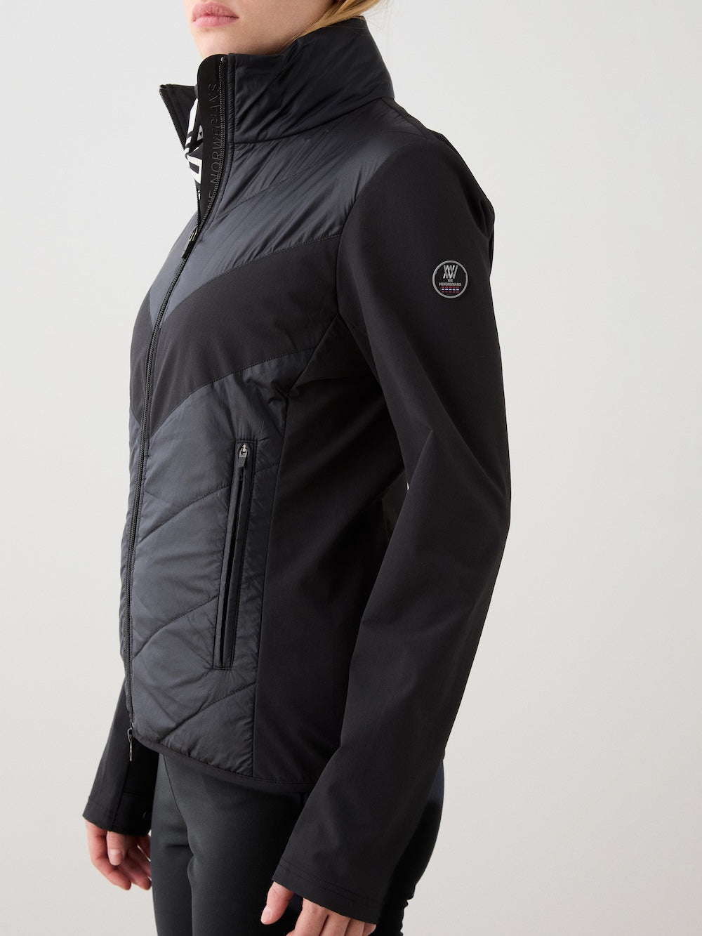 A person wears the We Norwegians Beito Jacket Women—a black, water-repellent zip-up with high collar, side pockets, and a logo patch on the upper left sleeve—shown from the side against a plain background.