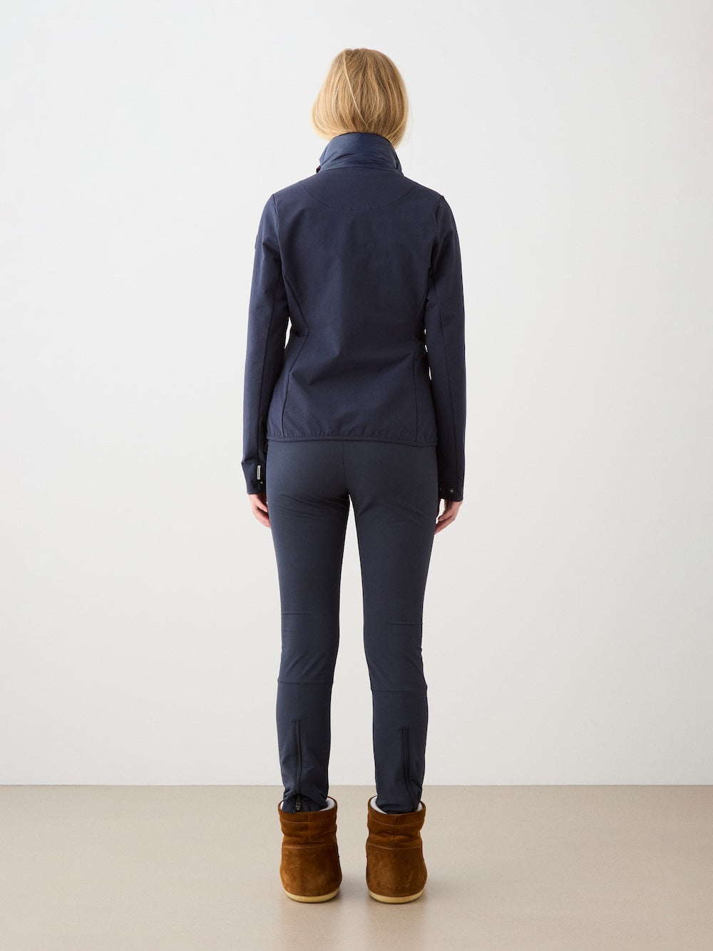 A person with light brown hair, seen from behind in a plain light-colored setting, wears the We Norwegians Beito Jacket Women in navy with matching pants and brown ankle boots.